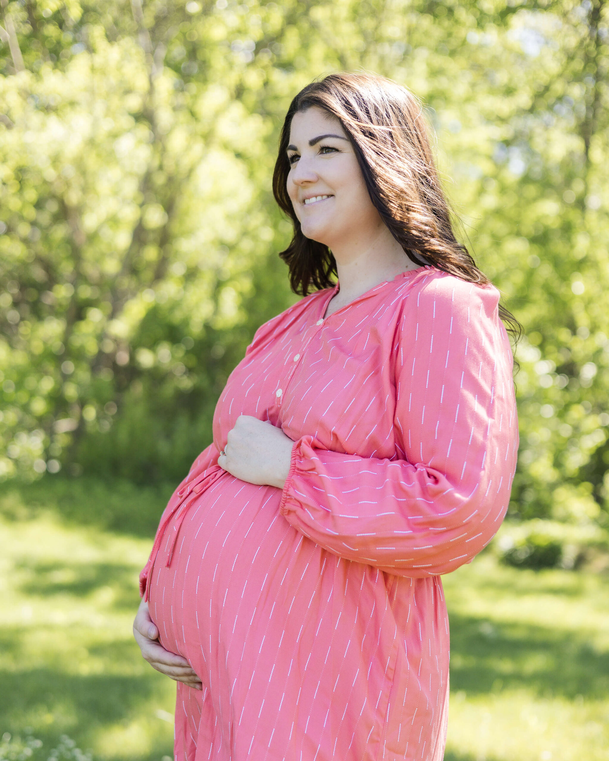 A happy expecting mother in a pink maternity dress smiles while standing in a park with hands on her bump