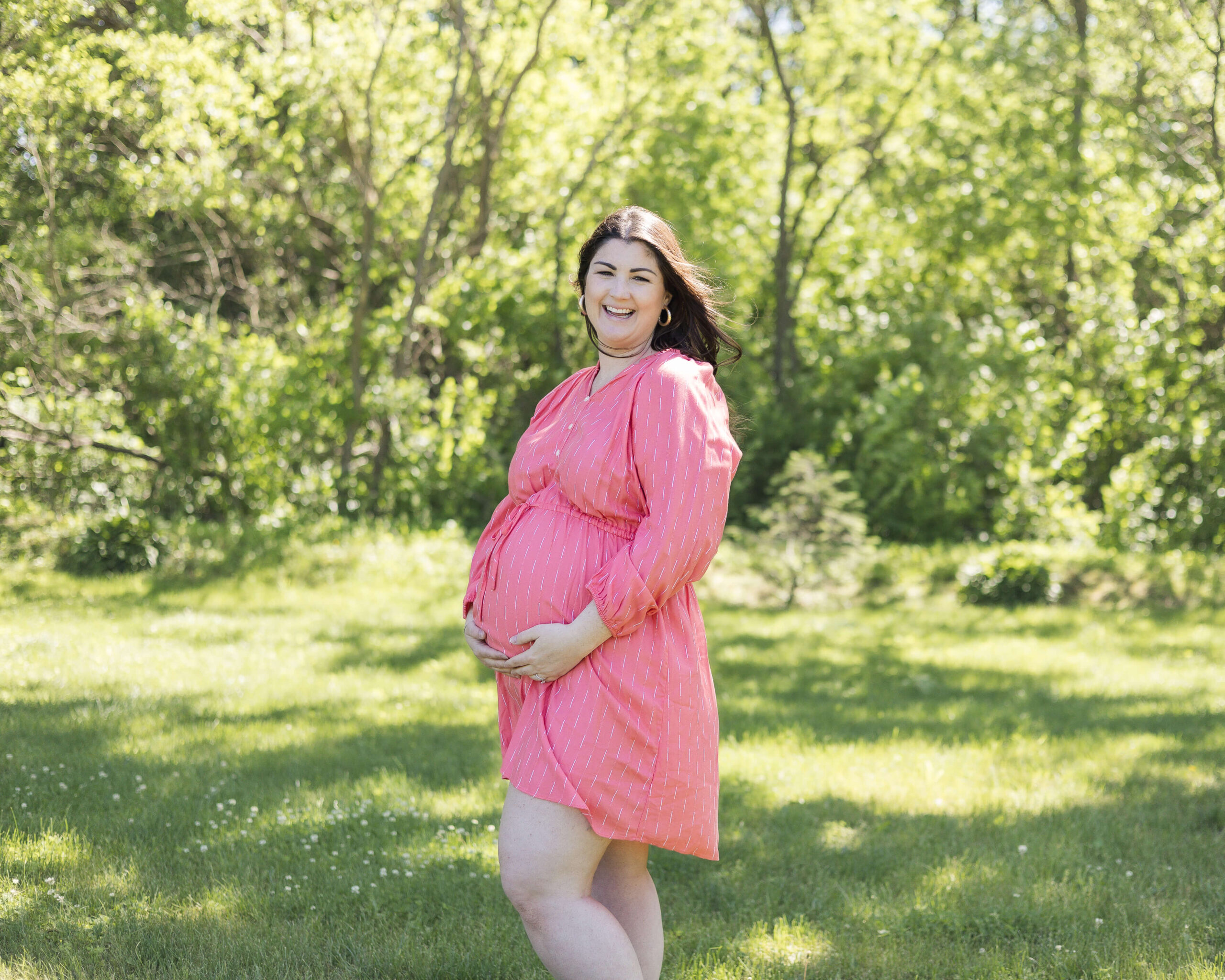 A laughing pregnant woman stands with hands under the bump in a pink maternity dress in a park after visiting birthing centers in Springfield, IL