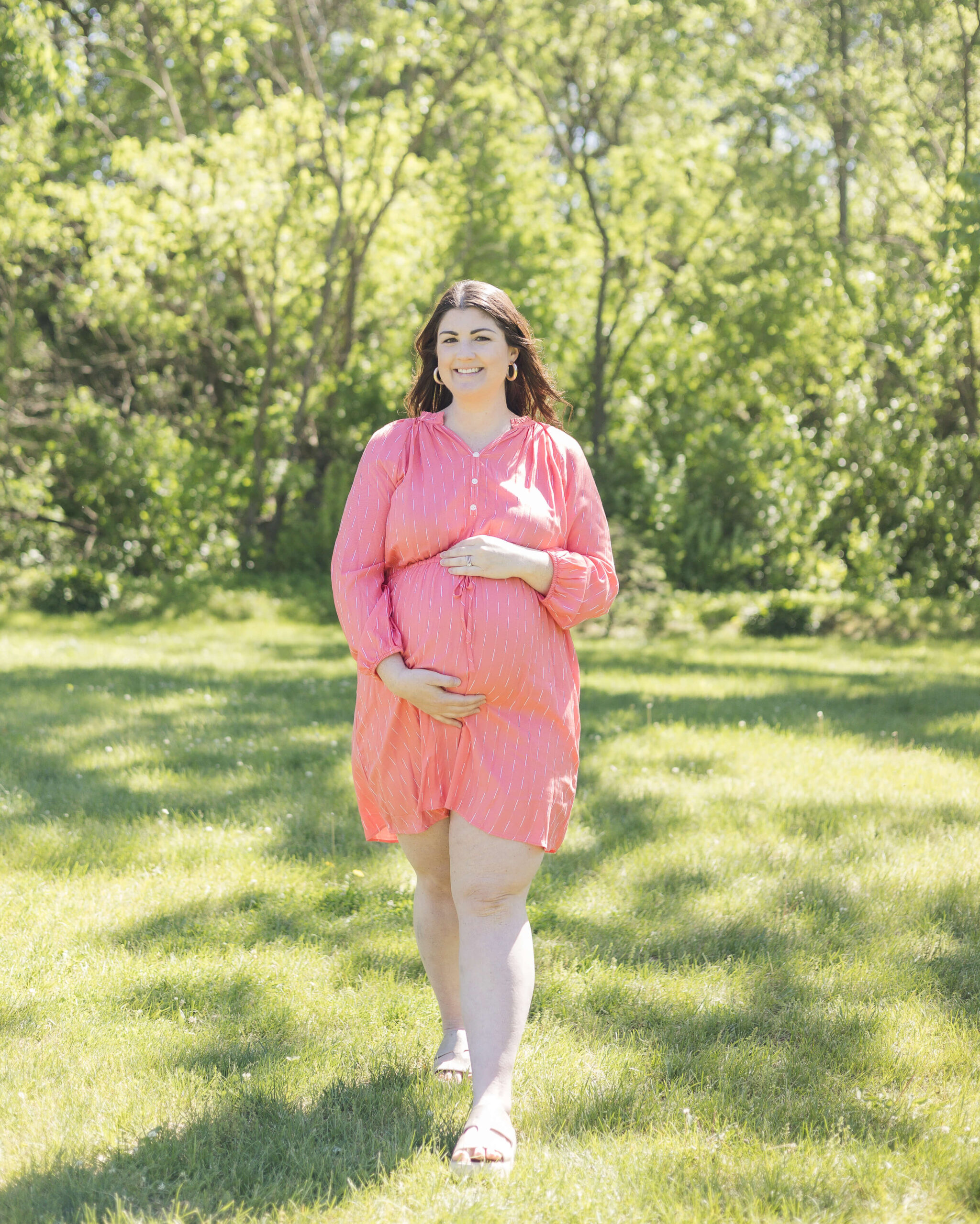 A smiling mother to be in a pink maternity gown walks in a park holding her bump after visiting birthing centers in Springfield, IL