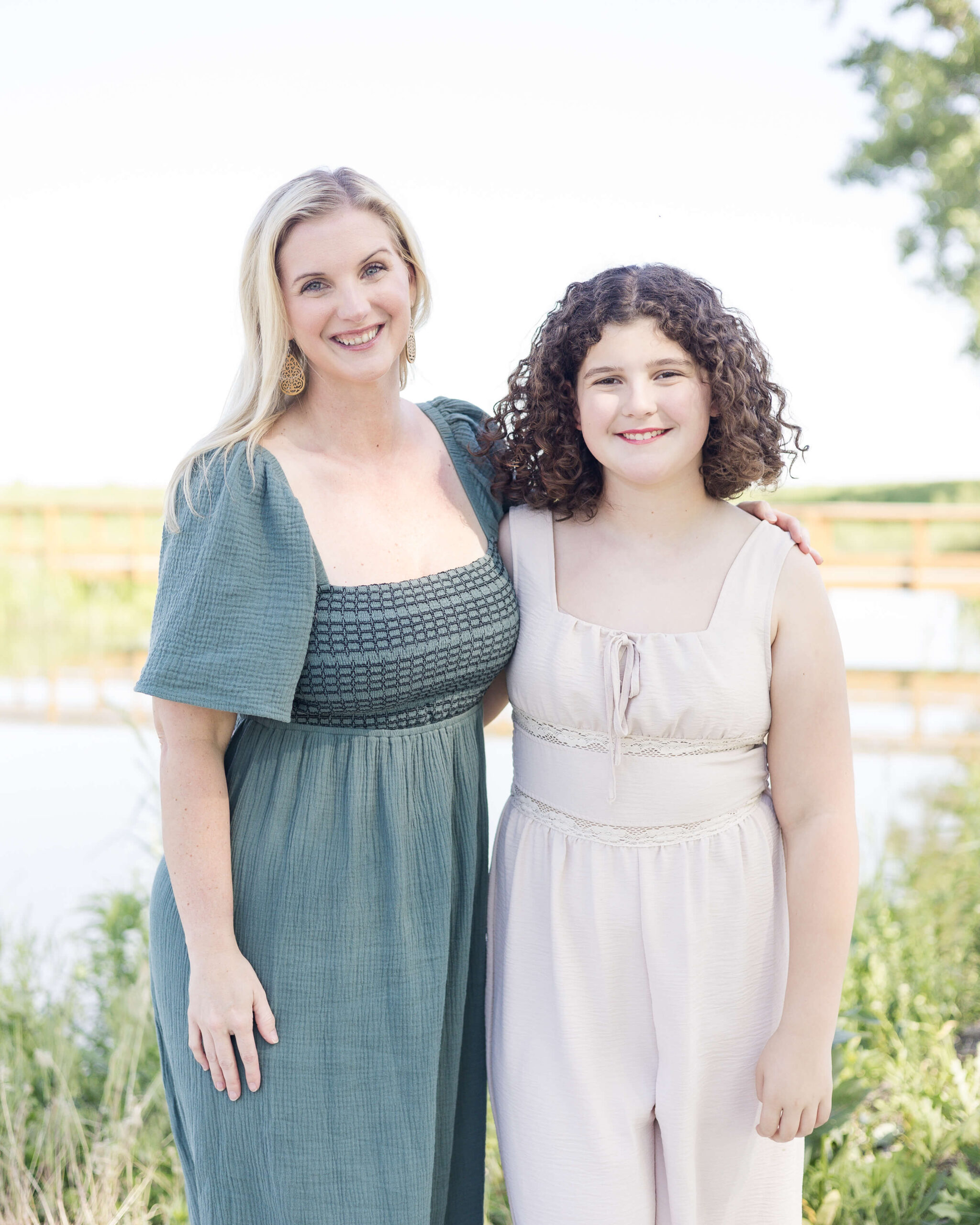 A smiling mother in a blue dress stands with her teen daughter in a pink romper by a lake