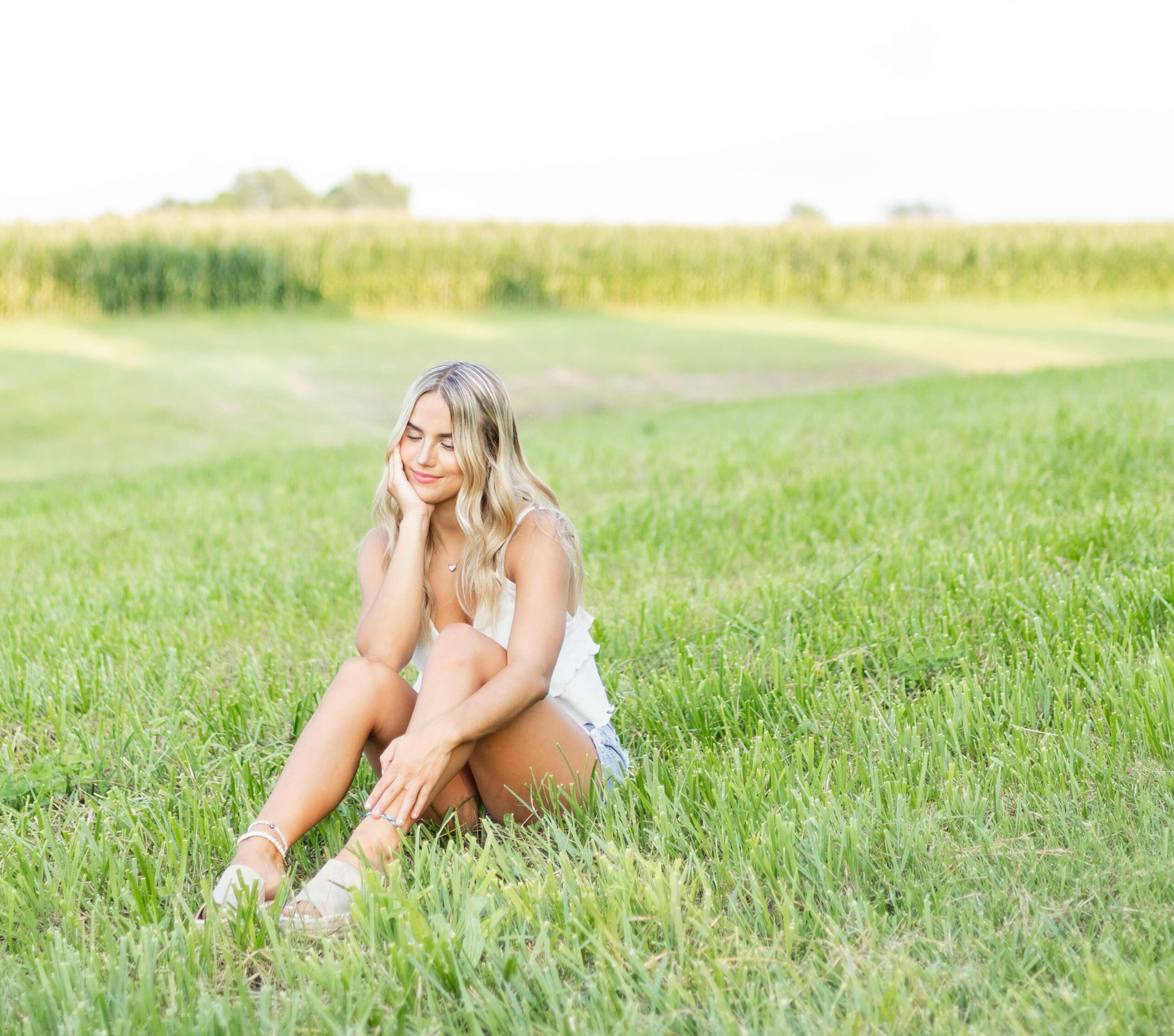 A high school senior in a white blows lounges in a field of grass by a corn field at sunset after visiting boutiques in Springfield, IL