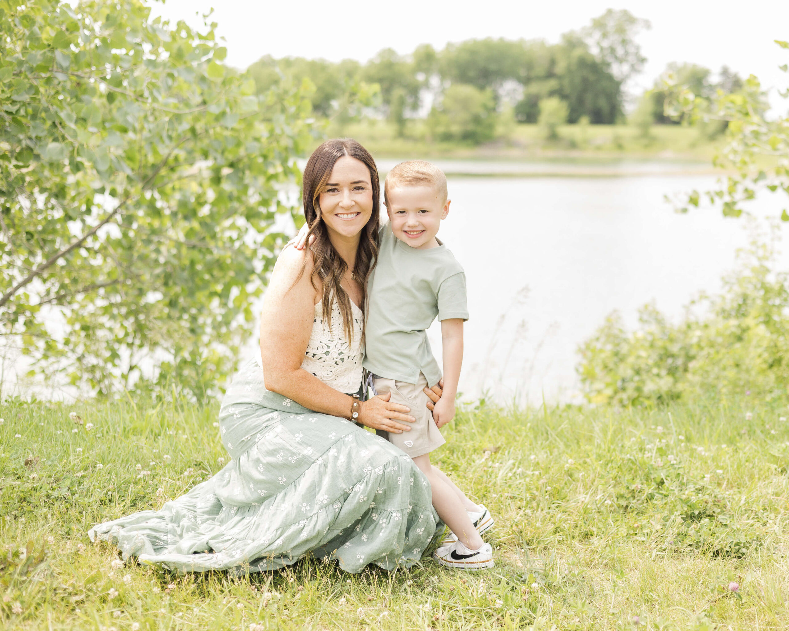 A smiling mother in a green dress kneels for a hug with her toddler son in a matching shirt by a lake after visiting boutiques in Springfield, IL