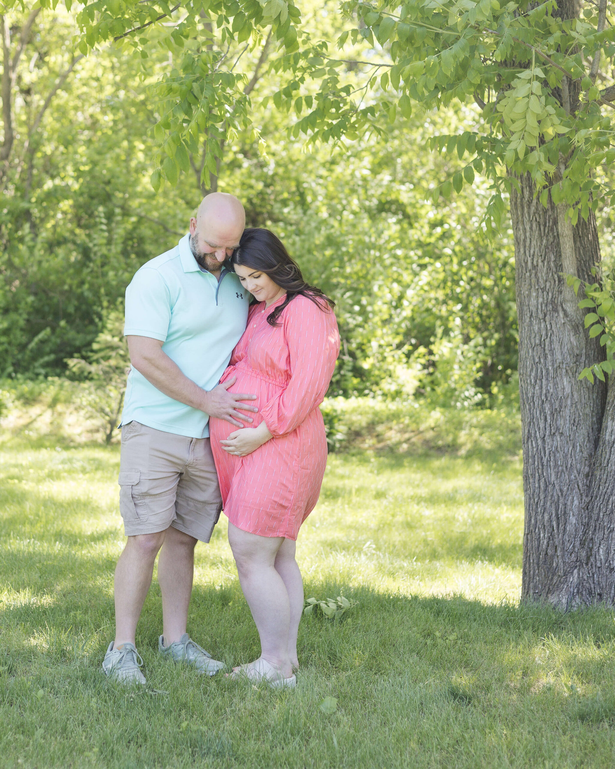 A happy expecting couple smile down at their hands on the bump as they snuggle under a tree in a park in blue and pink after meeting doulas in Springfield, IL