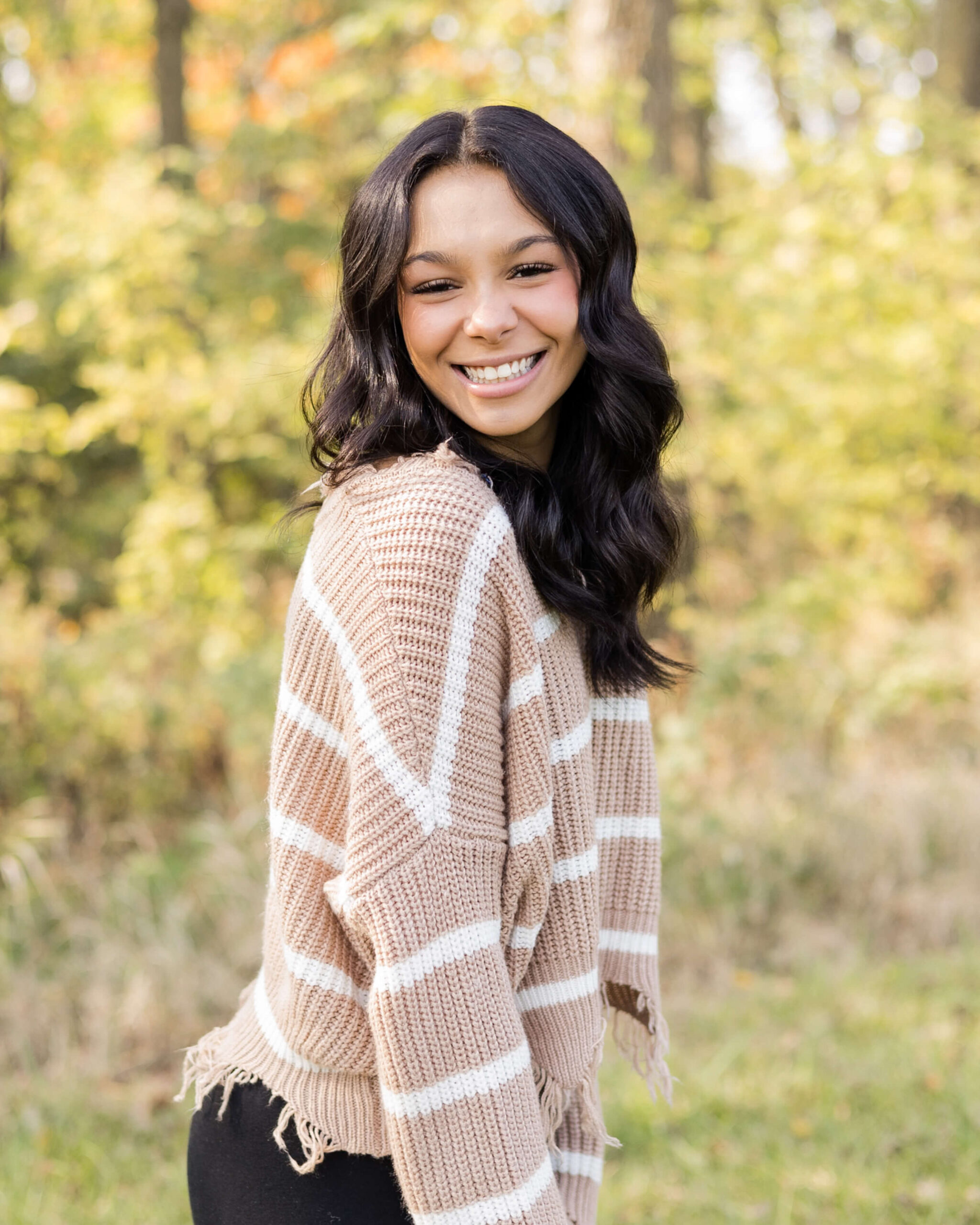 A woman smiles over her shoulder while walking a park at sunset in a brown sweater