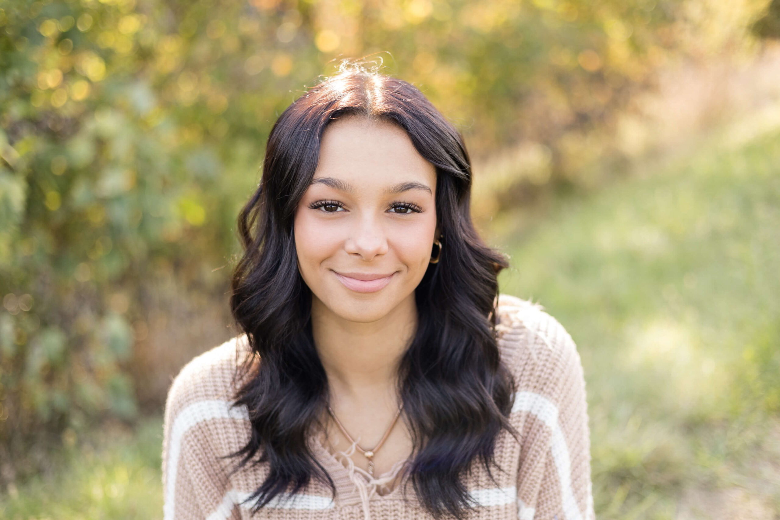 Closeup of a smiling high school senior with dark wavy hair in a brown sweater in a park at sunset after finding hair salons in Springfield, IL