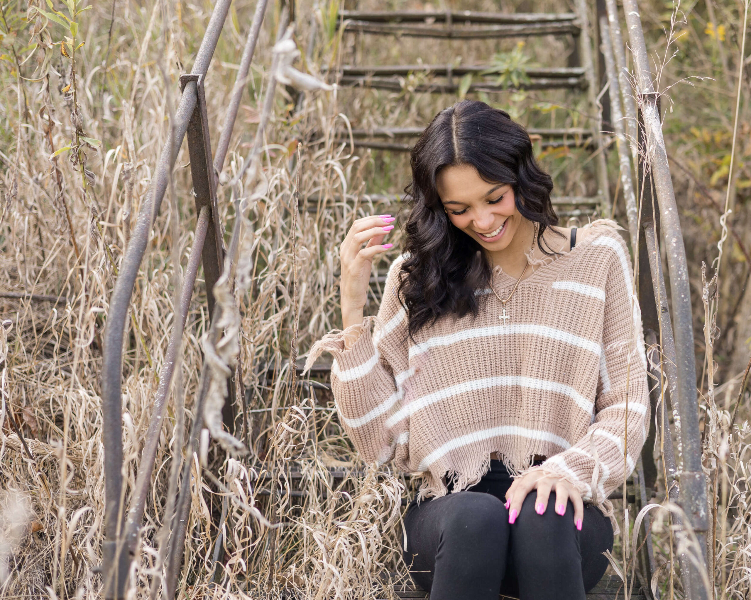 A high school senior sits on a metal stair set in a field of overgrown grass in a brown sweater laughing after finding hair salons in Springfield, IL