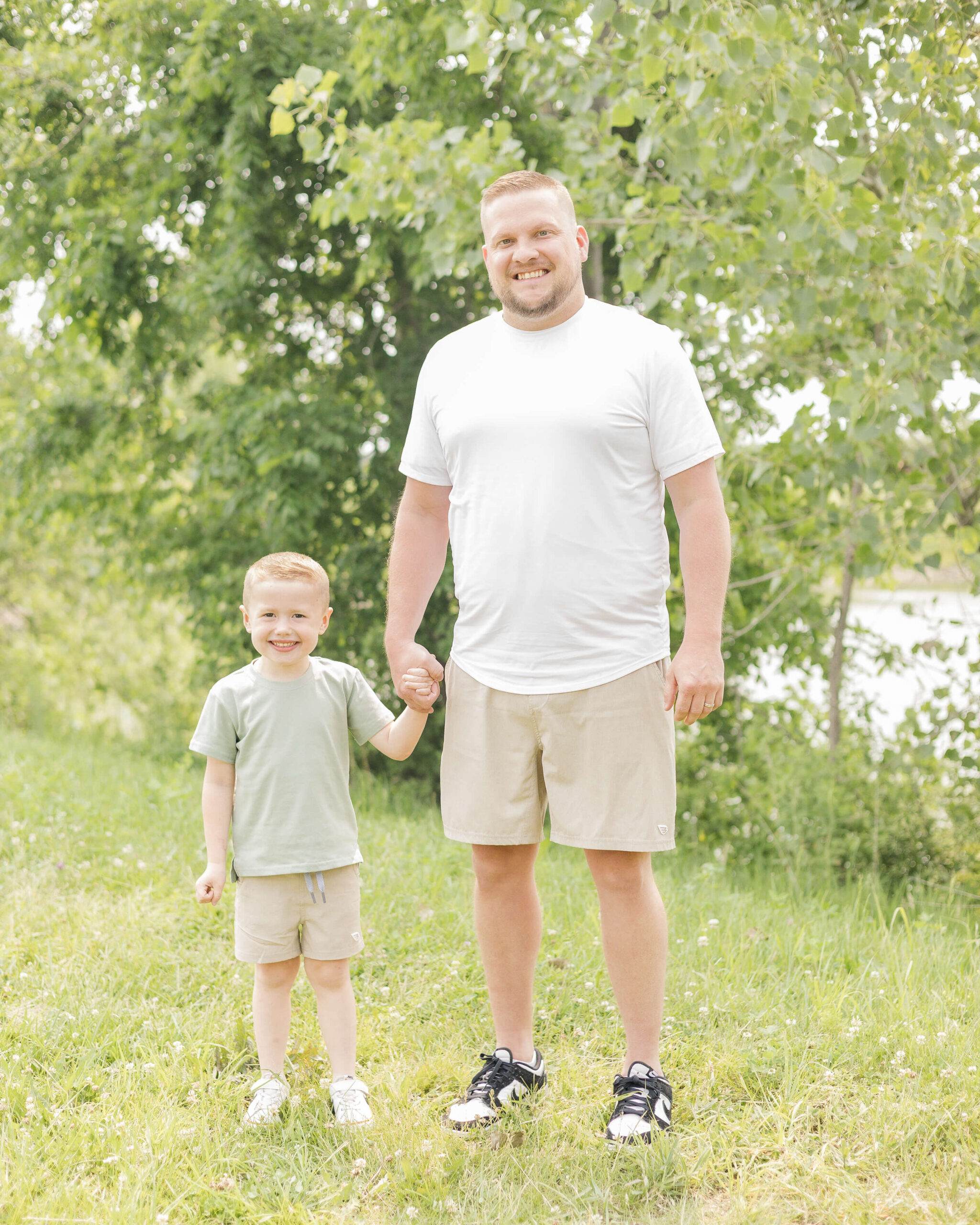 A happy dad in a white shirt stands holding his toddler son's hand in a green shirt by a lake after finding kid-friendly things to do in Springfield, IL