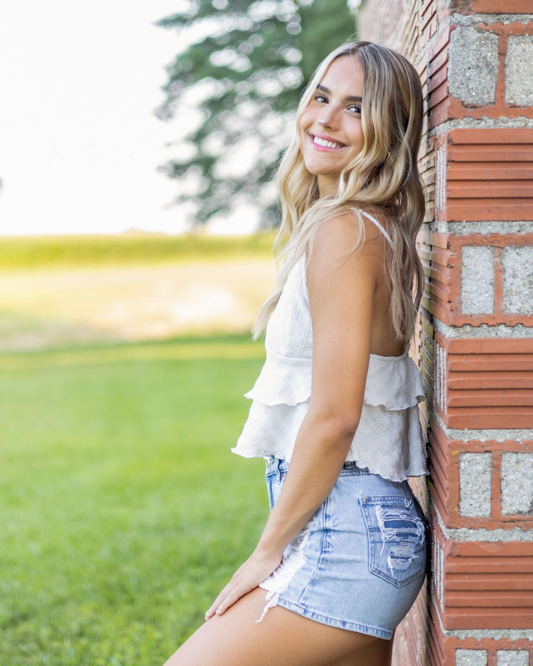 A smiling blonde high school senior in a white shirt leans on a brick wall smiling over her shoulder