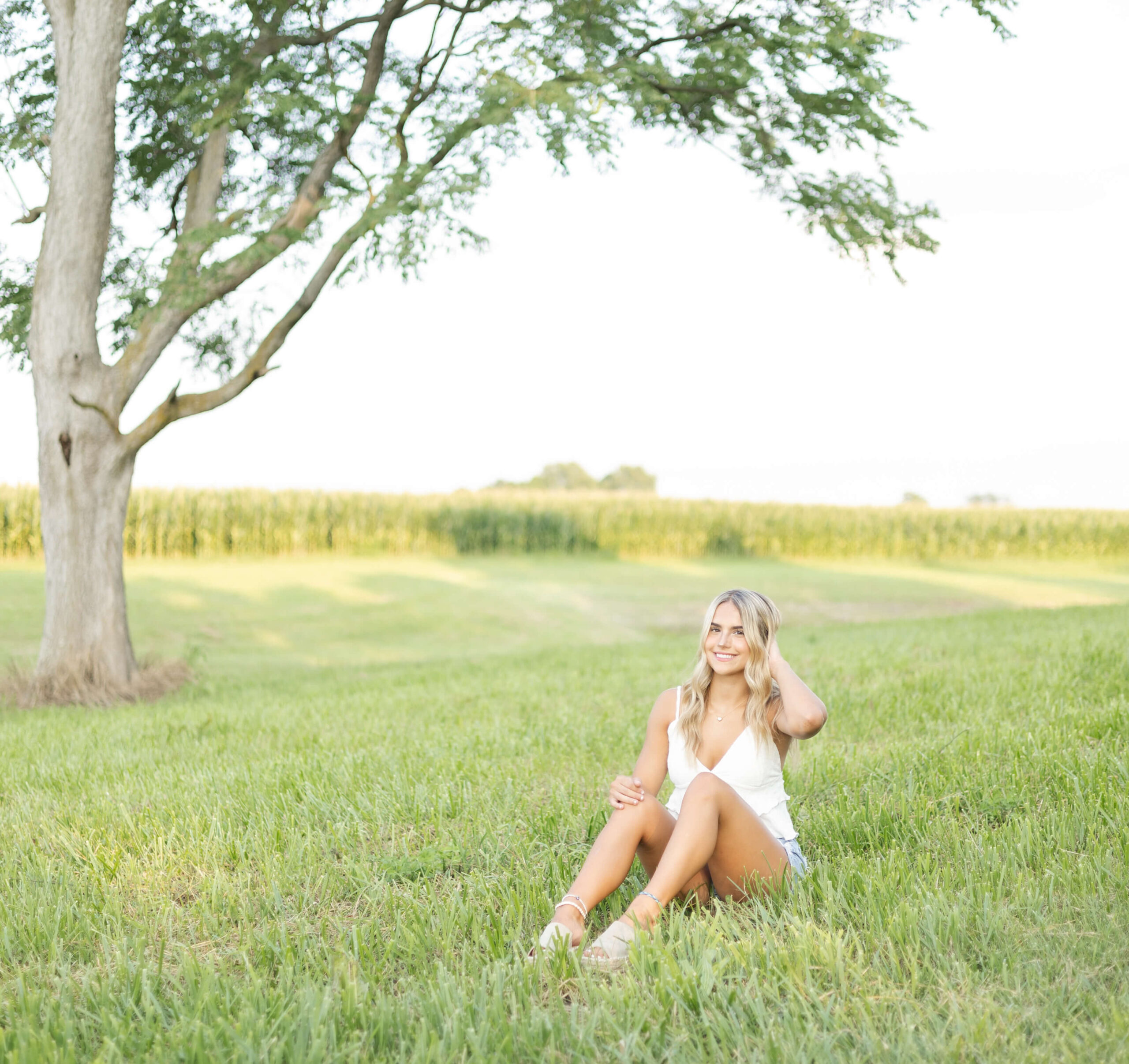 A high school senior in a white shirt and shorts sits in grass by a corn field at sunset after finding makeup artists in Springfield, IL