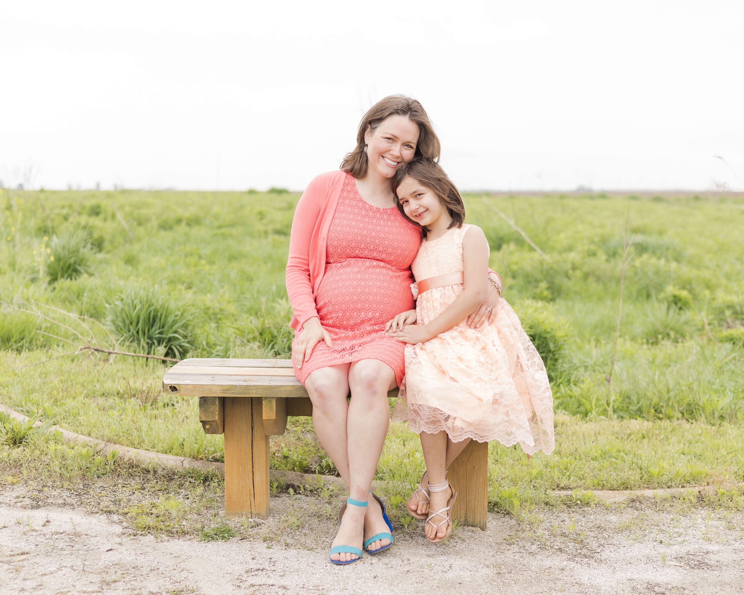 A smiling pregnant mother sits on a park bench hugging her toddler daughter in a peach dress