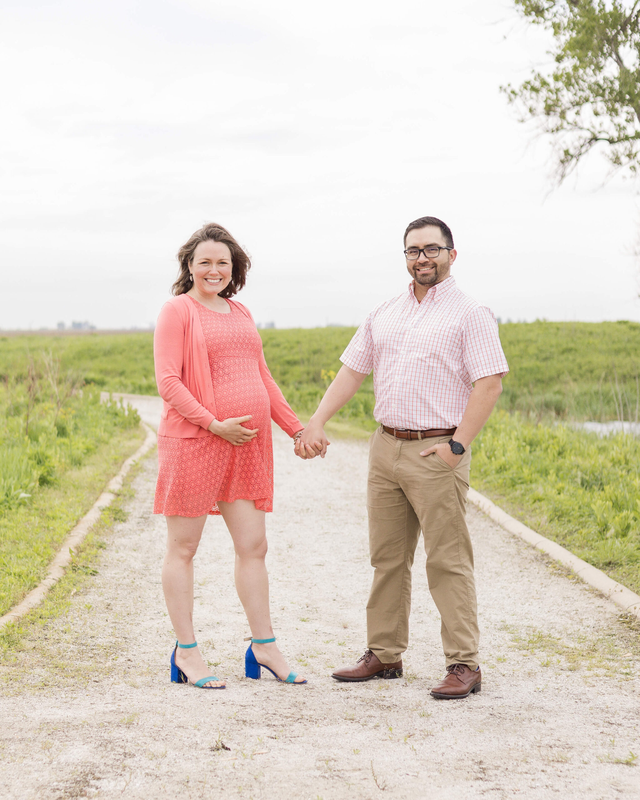 A pregnant woman in a pink maternity dress smiles while holding hands with her husband in a pink shirt in a park trail after finding a midwife in Springfield, IL