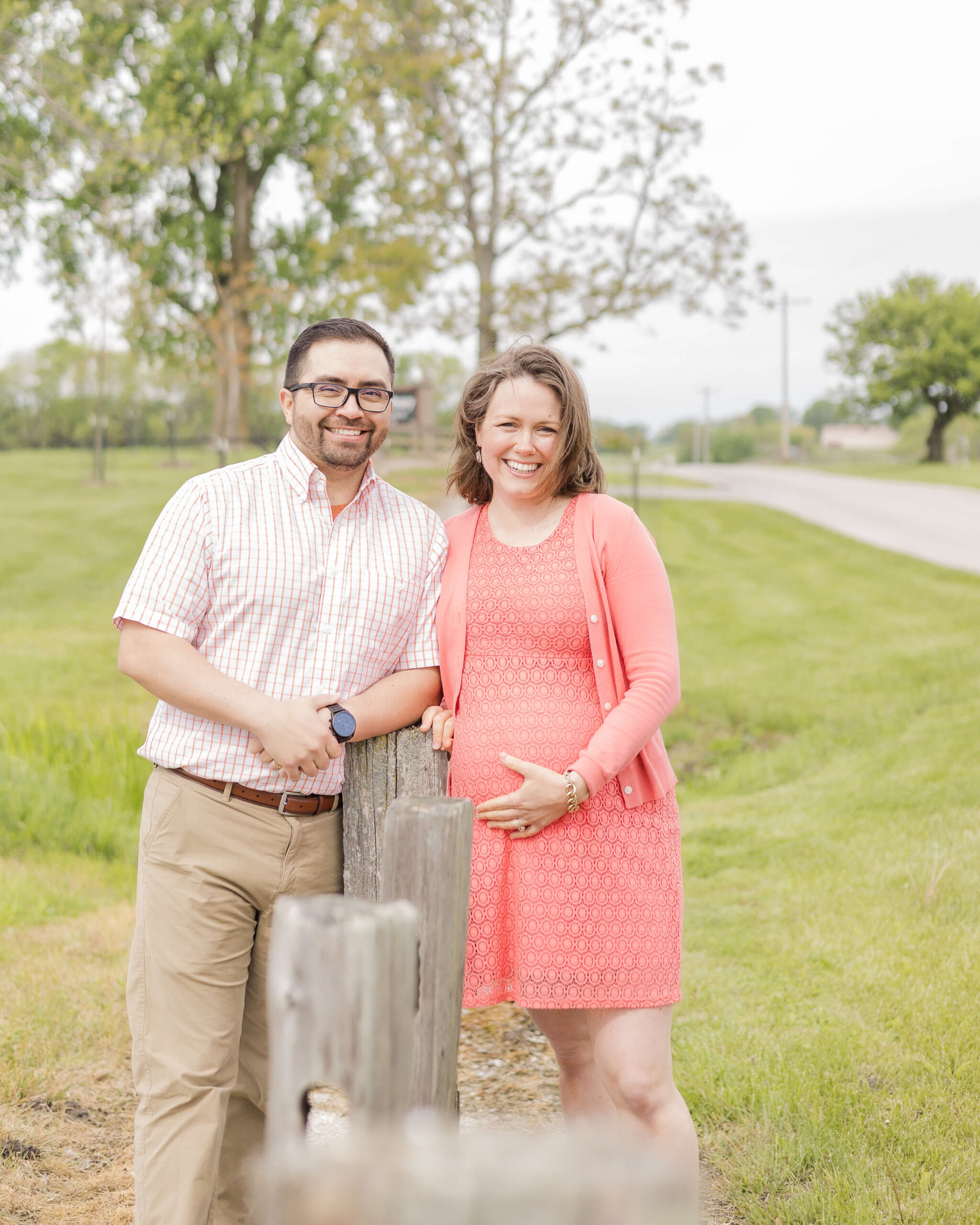 Happy expecting parents in pink smile while leaning on wooden fence post in a park after finding a midwife in Springfield, IL