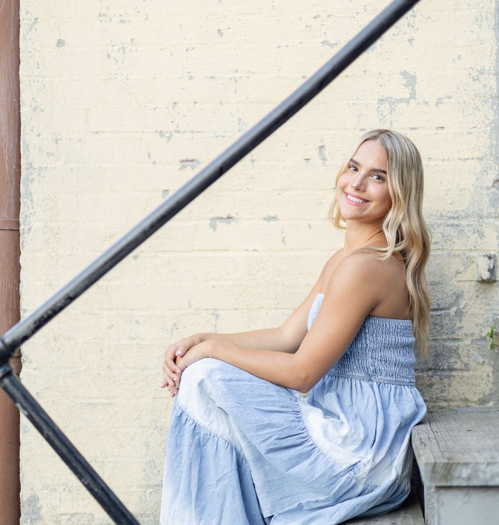 A smiling high school senior in a blue dress sits on rustic alley stairs smiling over her shoulder