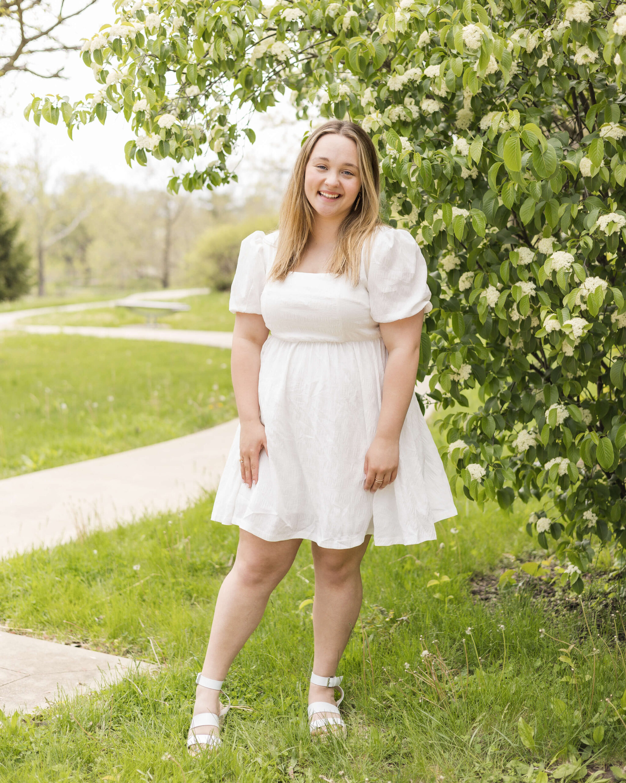 A happy high school senior in a white dress smiles while standing by a flowering bush after finding prom dresses in Springfield, IL