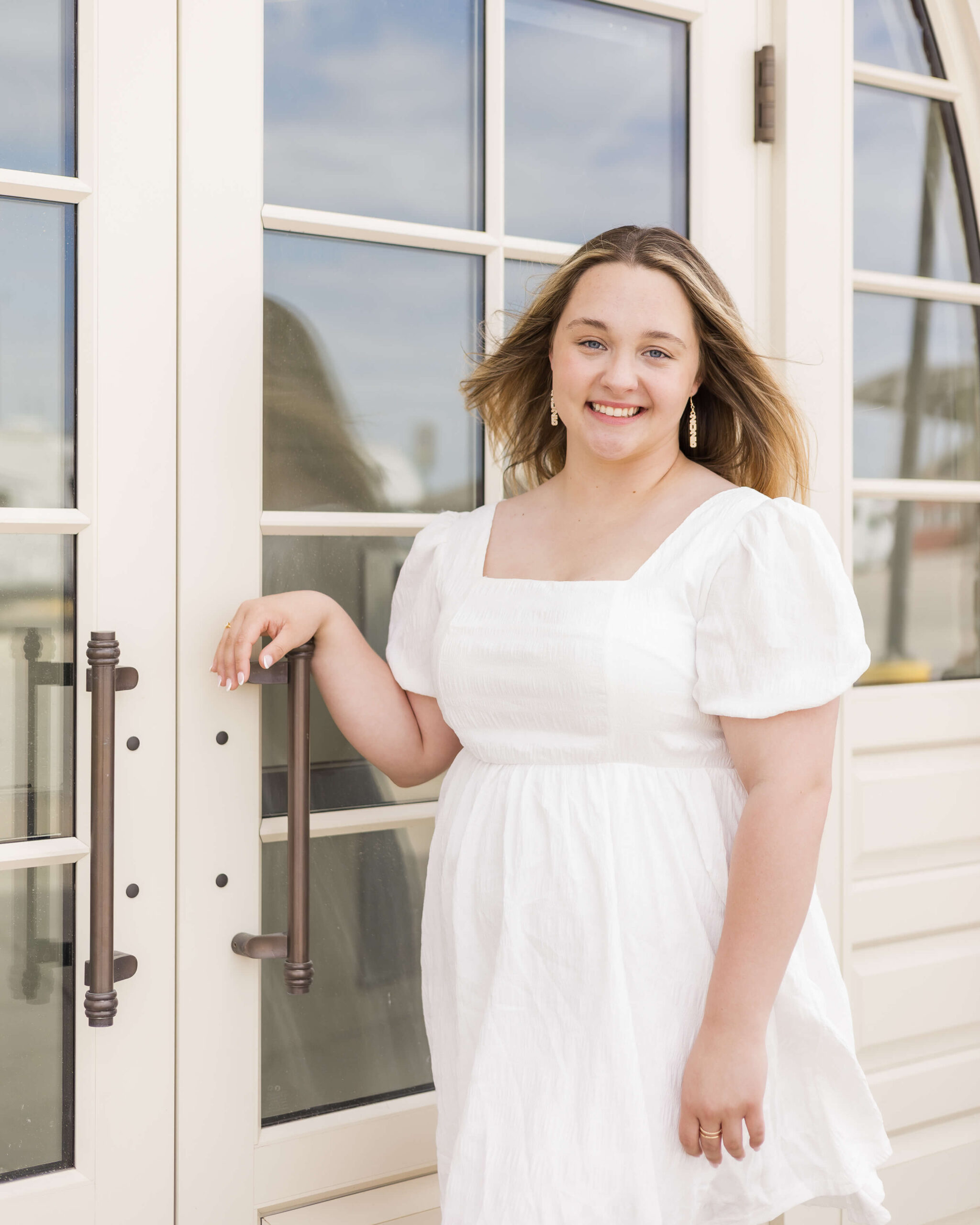 A high school senior in a white dress smiles while leaning on a door handle on a porch after finding prom dresses in Springfield, IL
