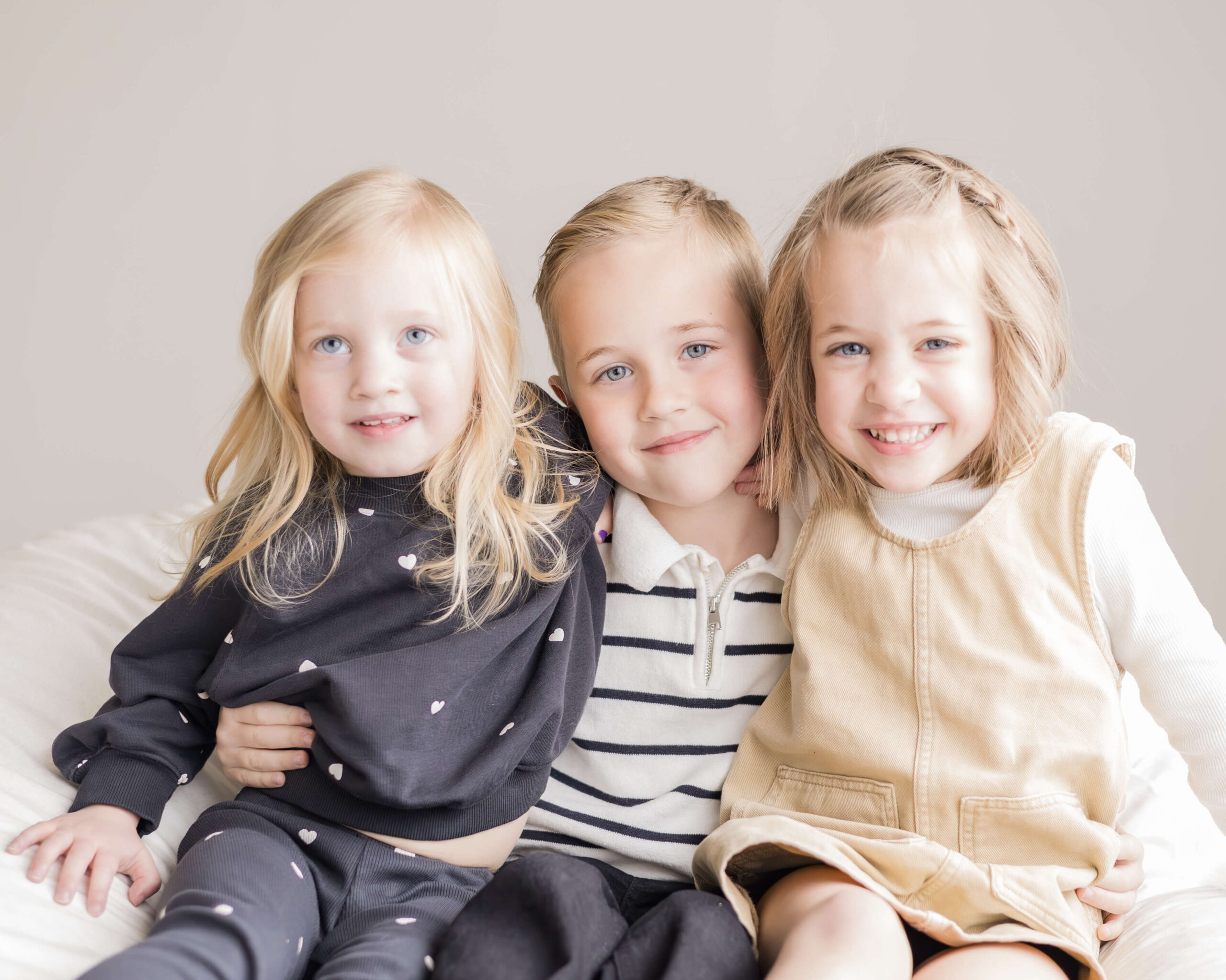 A young boy sits on a chair with his two younger sisters smiling in his lap with arms around each other