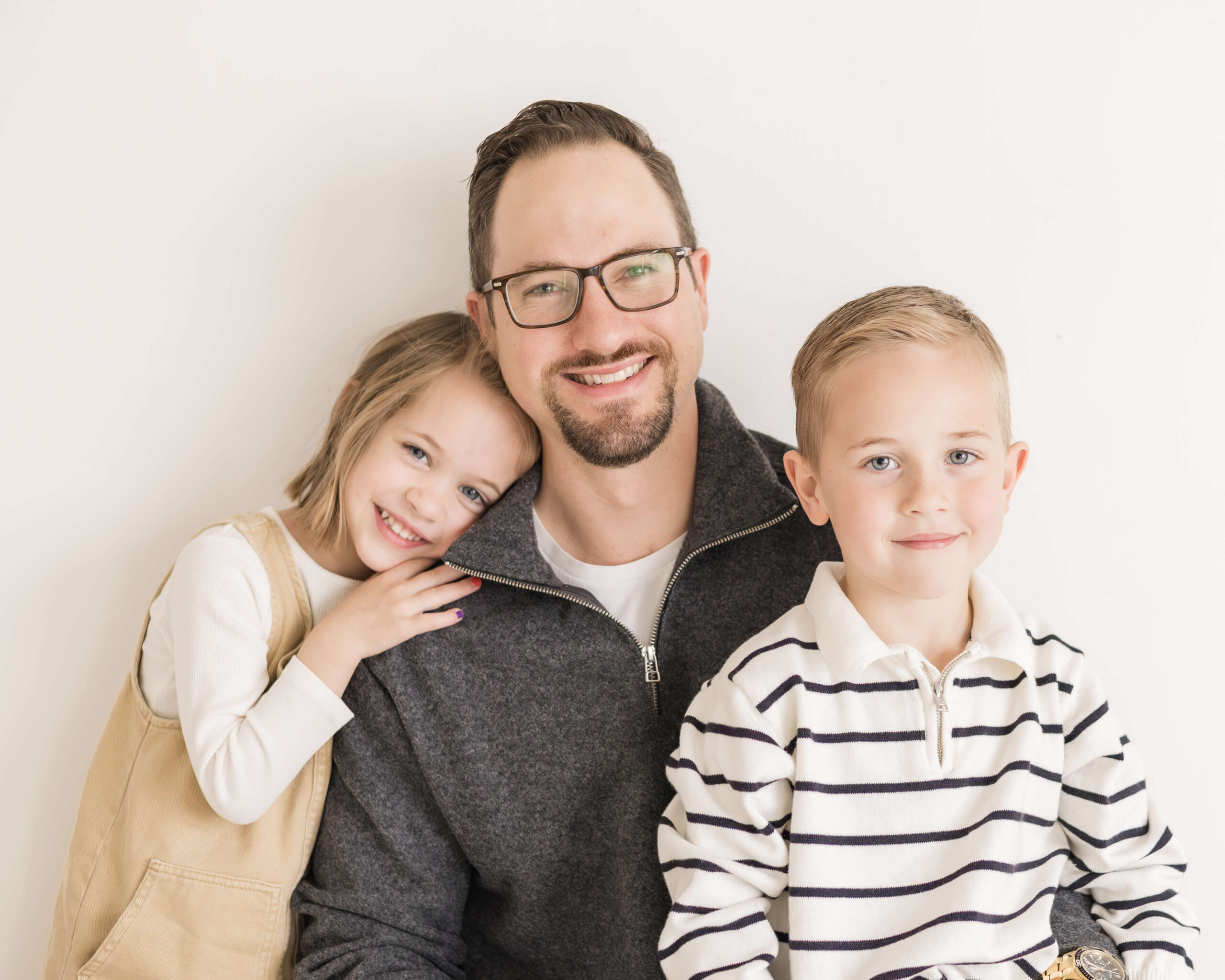 A happy dad in a grey sweater smiles while sitting with his toddler son and daughter cuddling him and sitting in his lap before visiting toy stores in Springfield, IL