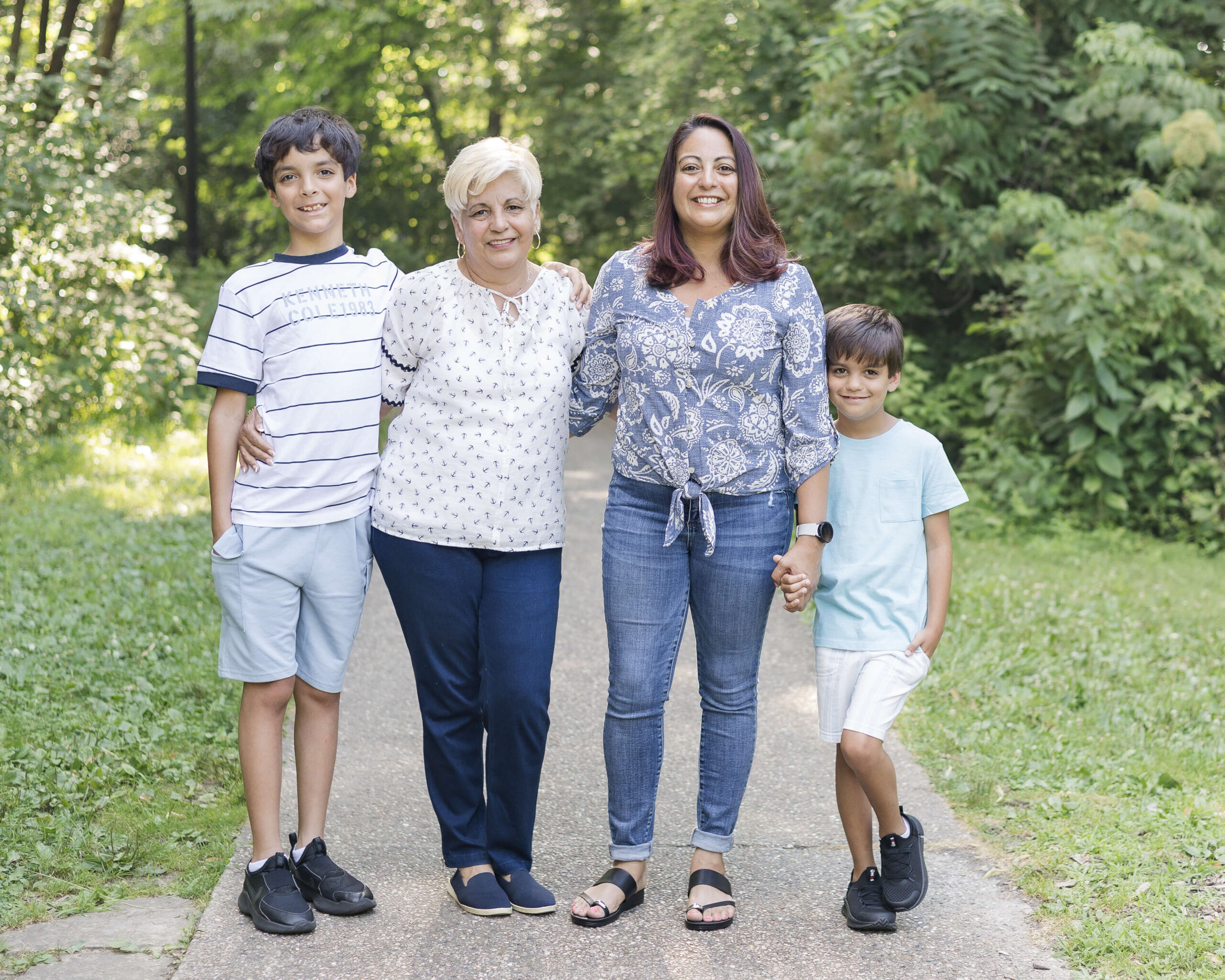 A mom and grandma stand smiling in a park sidewalk holding hands with two young brothers