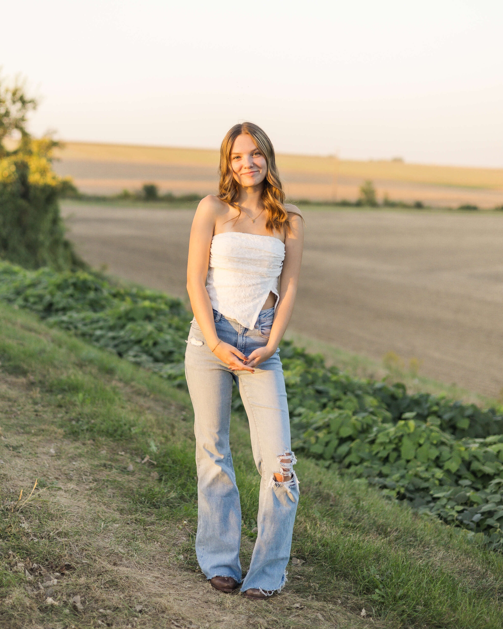 A smiling teenage girl in a white blouse and jeans walks a trail overlooking a farm at sunset