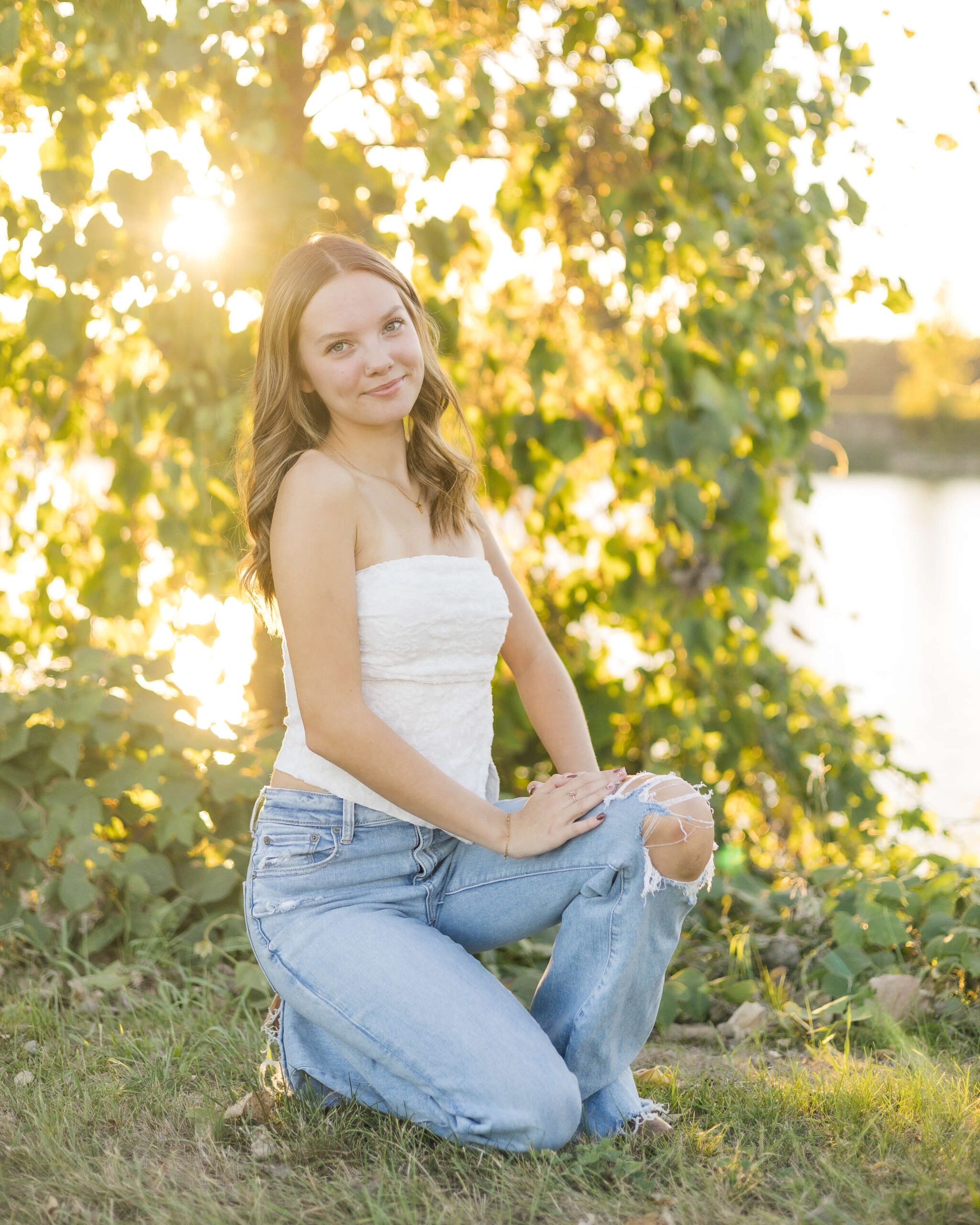 A high school senior in a white blouse and jeans kneels by a lake at sunset with a smile after visiting nail salons in springfield il