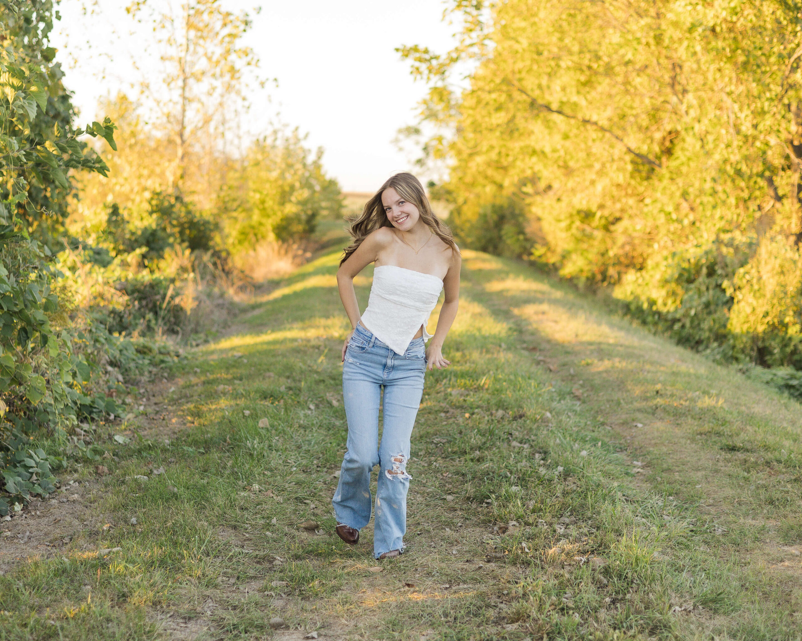 A high school senior in a white tube top nd jeans walks in a park trail at sunset after finding nail salons in springfield il