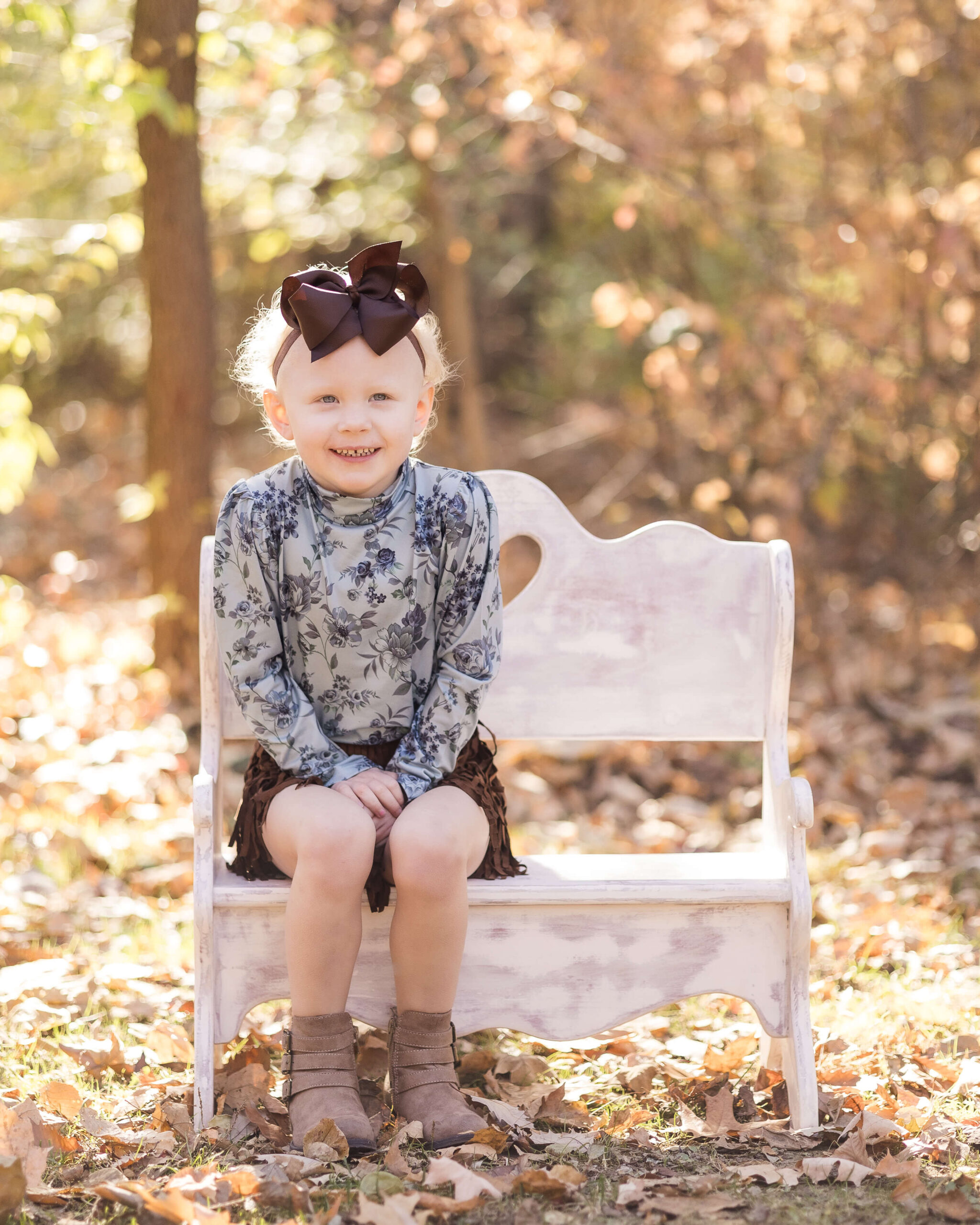 A toddler girl in a blue flower top and brown skirt sits on a wooden bench in the fall leaves with a smile and hands in lap after meeting pediatric dentists in springfield IL