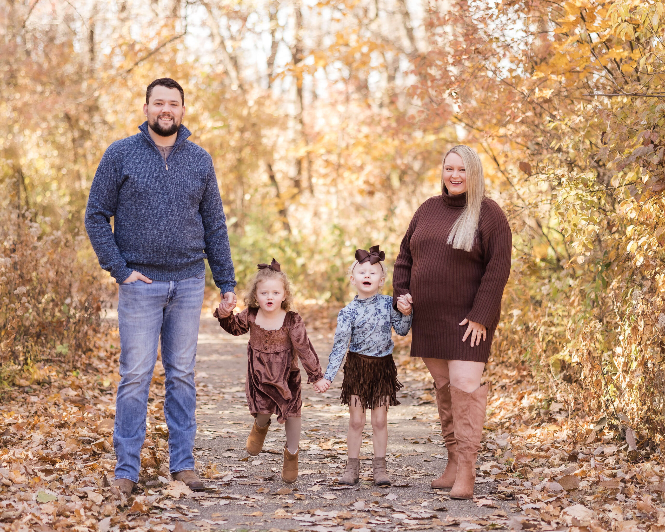 Happy mom and dad in brown and blue hold hands with their two toddler daughters while walking a trail in fall after finding pediatric dentists in springfield IL