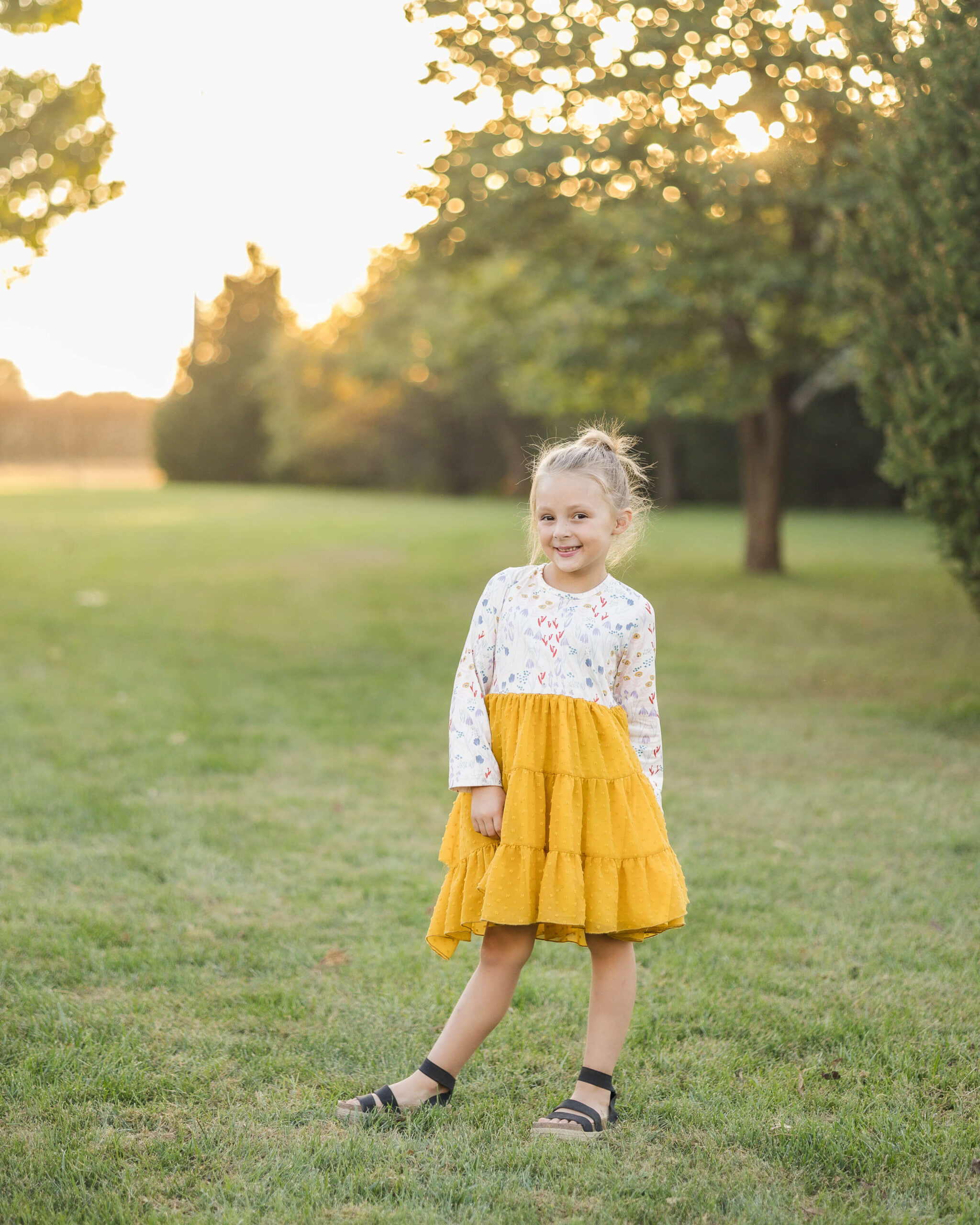 A toddler girl in a yellow and white dress stands in a park lawn at sunset with a big smile