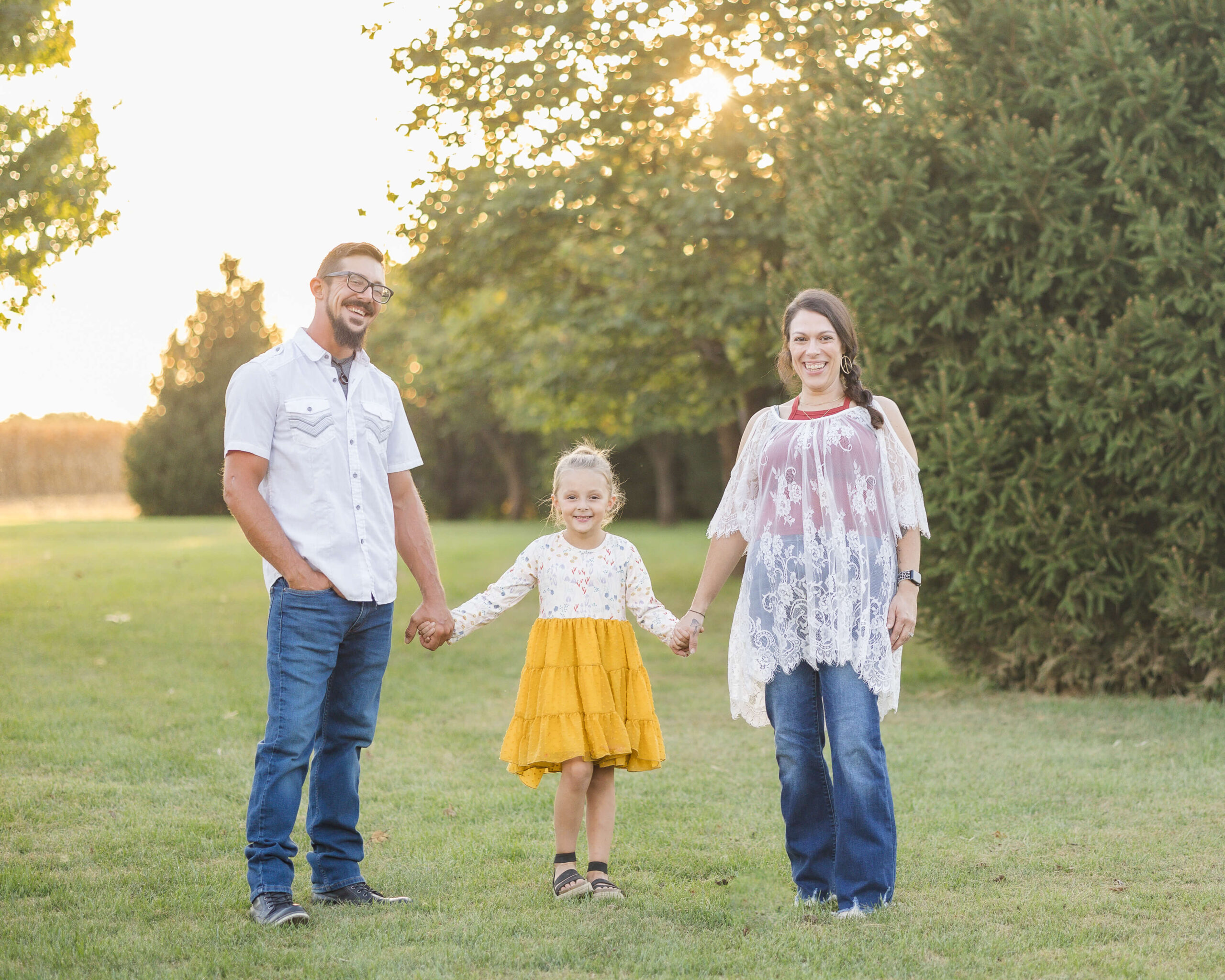 Happy mom and dad in white tops and jeans hold hands with their young daughter in a yellow dress after visiting pediatricians in springfield IL