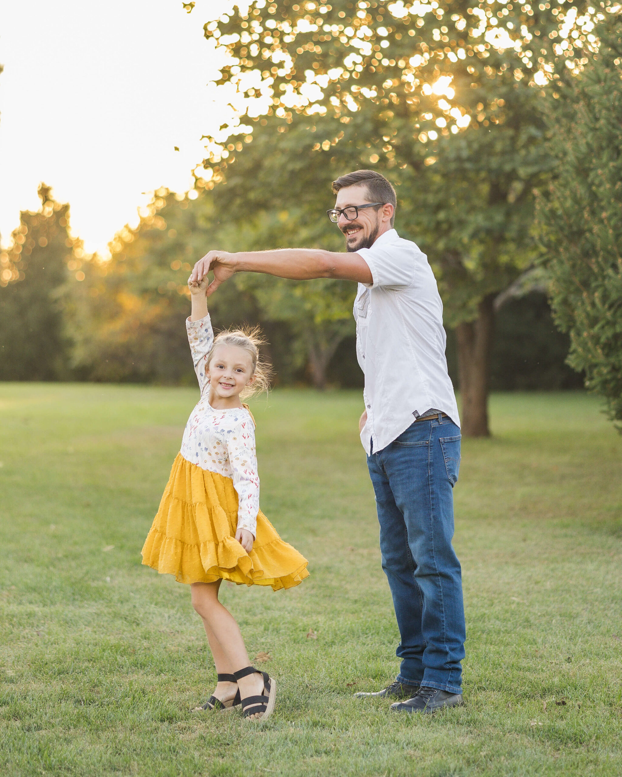 A toddler girl in a yellow and white dress twirls and dances holding dad's hand in a park lawn at sunset after finding pediatricians in springfield IL