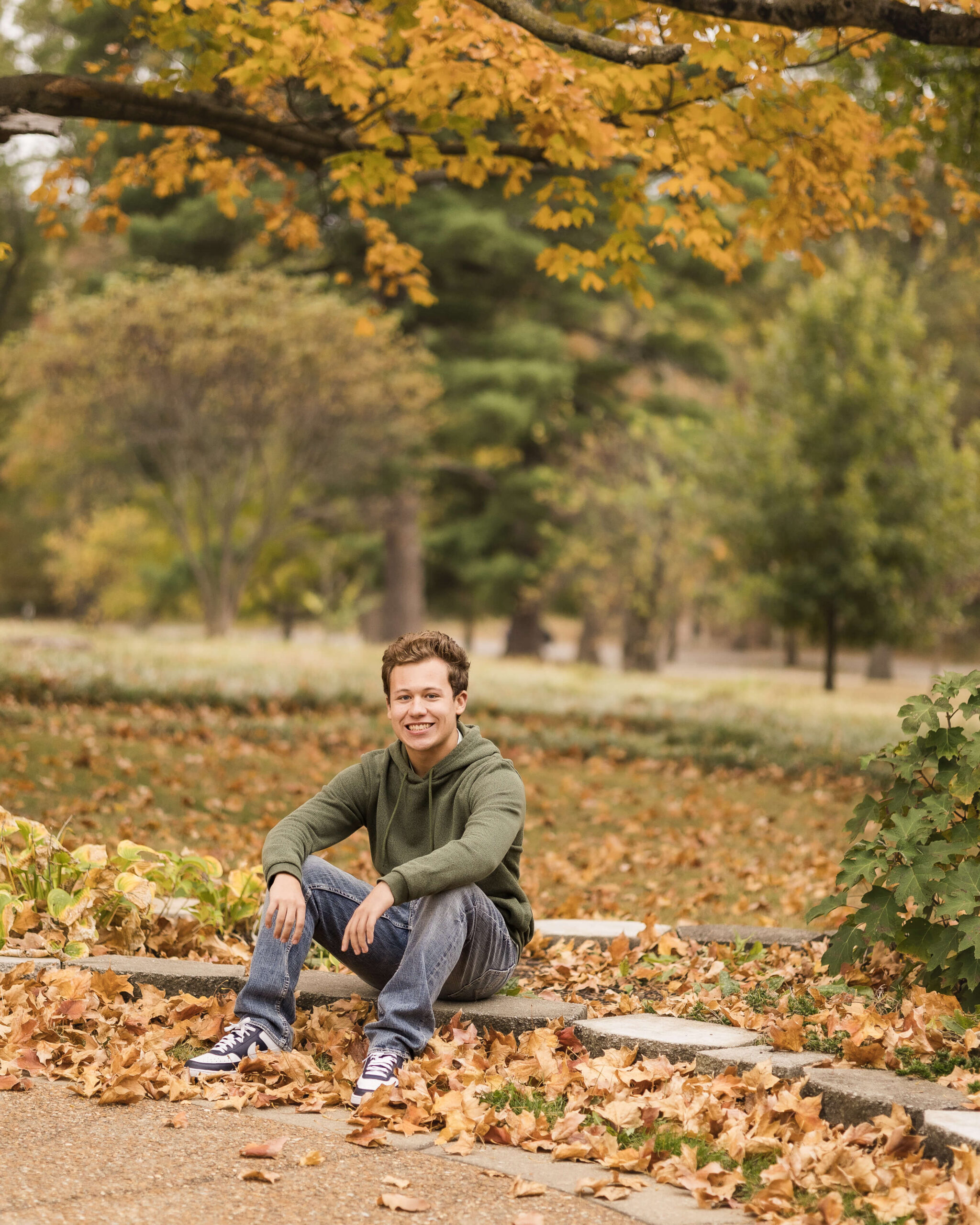 A happy teenage boy in a green hoodie sits on a garden wall in a park during fall