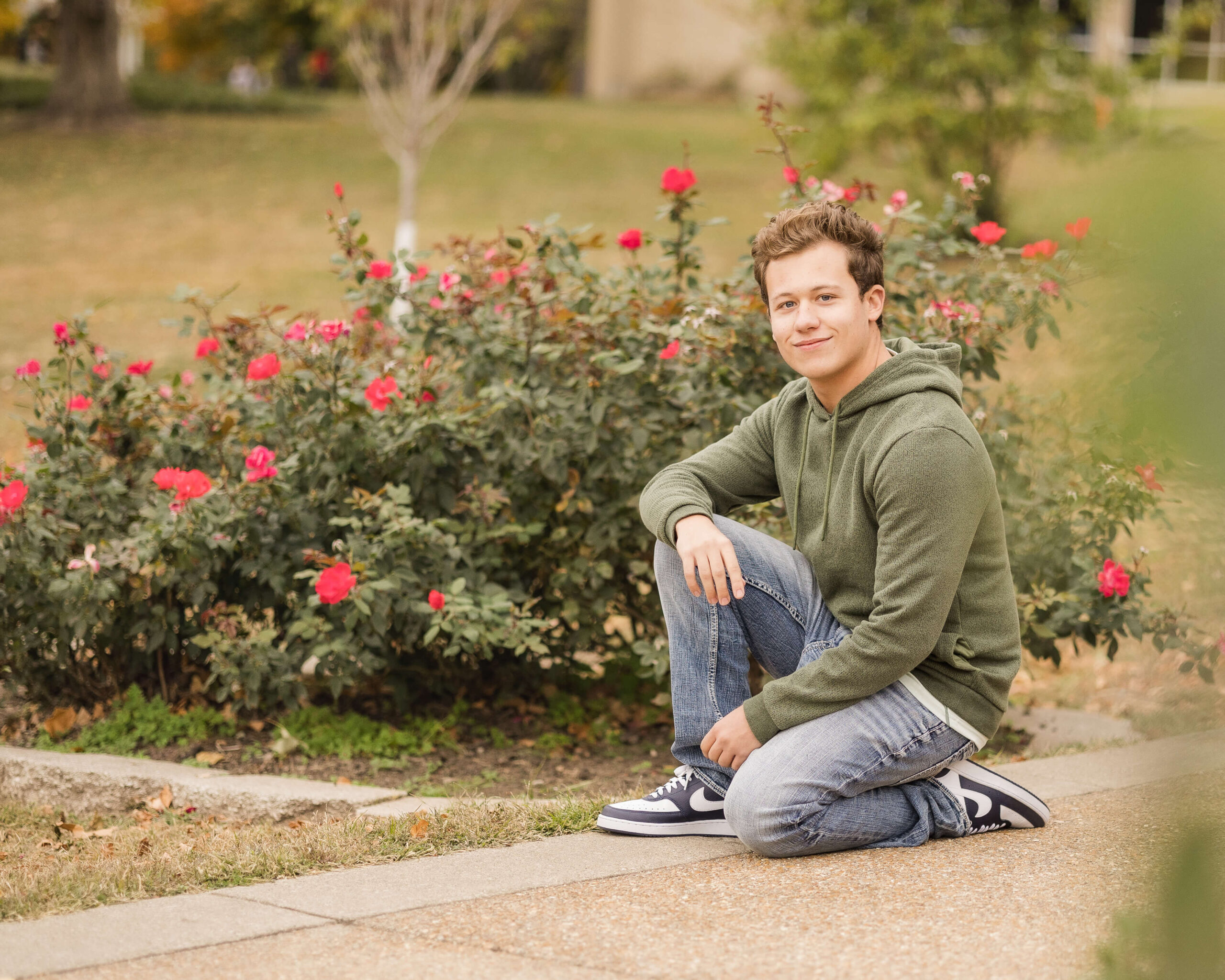 A high school senior kneels by some roses in a park in jeans and green hoodie while smiling after finding tuxedo rental in springfield il