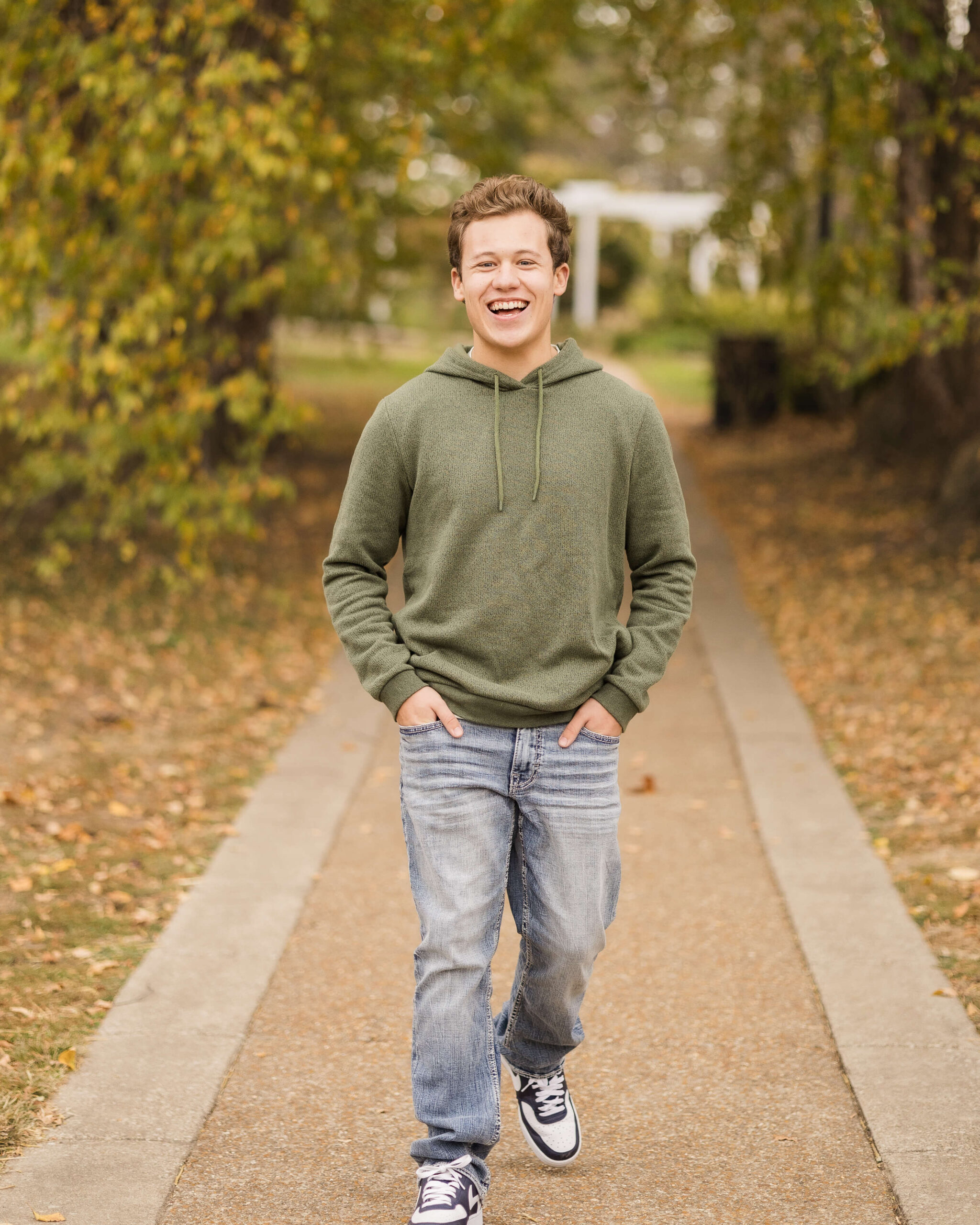 A high school senior walks a park sidewalk with a big smile and hands in jean pockets in a green hoodie after finding tuxedo rental in springfield il