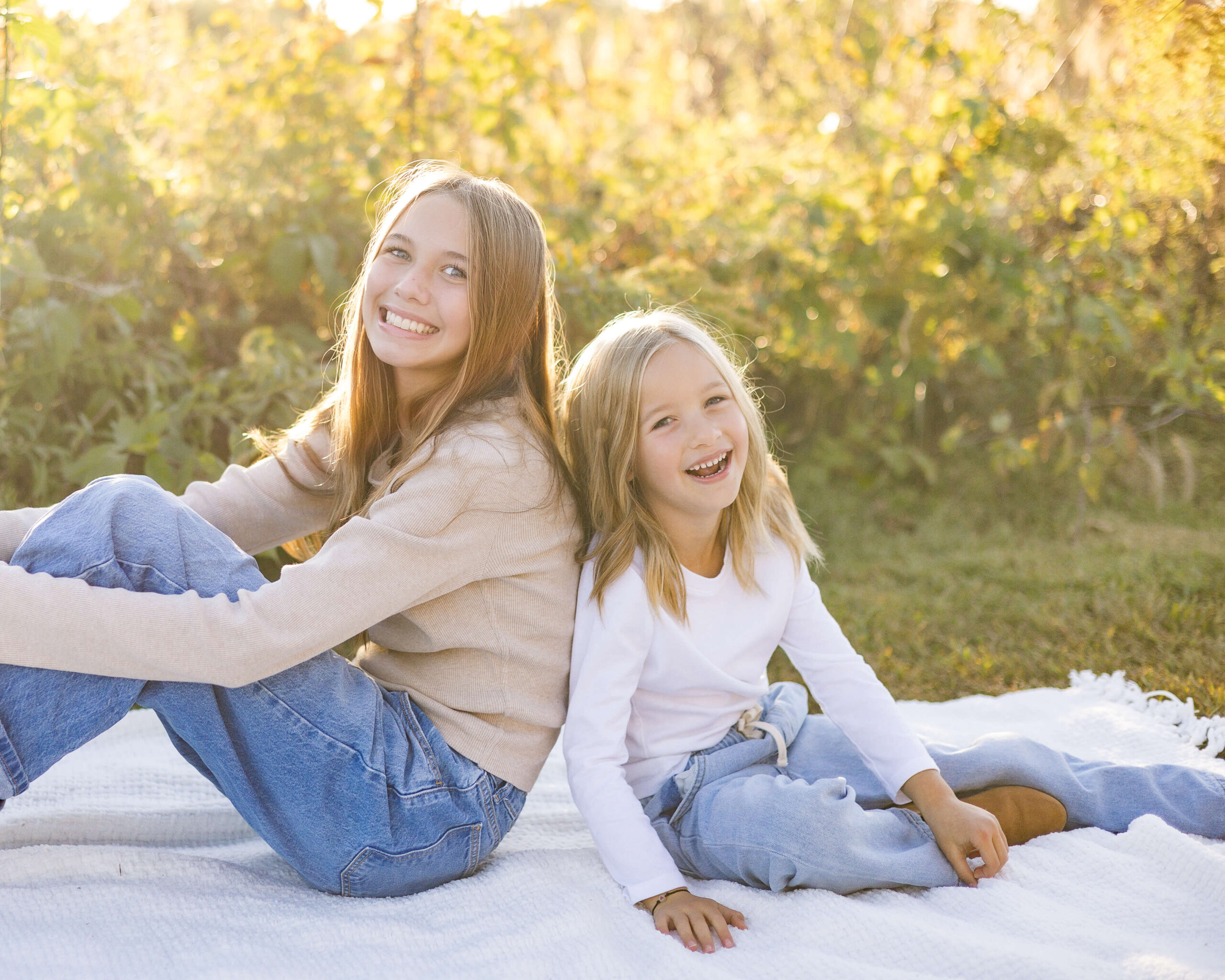 Smiling young sisters in jeans and long sleeve shirts laugh while sitting back to back on a picnic blanket after enjoying churches in springfield il