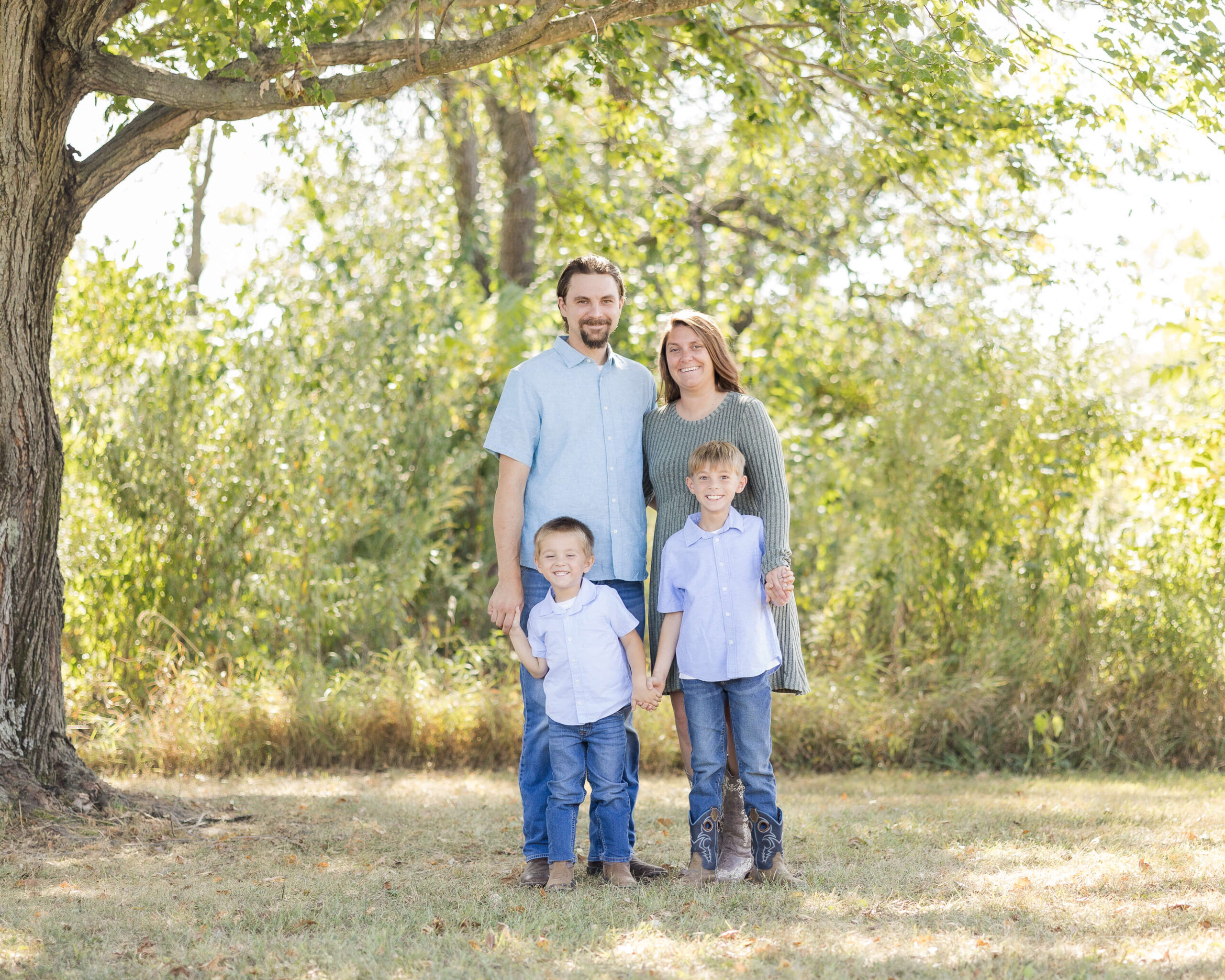 Happy mom and dad stand under a tree in a park with their two giggling toddler sons in matching jeans and blue shirts before heading to county fairs in illinois