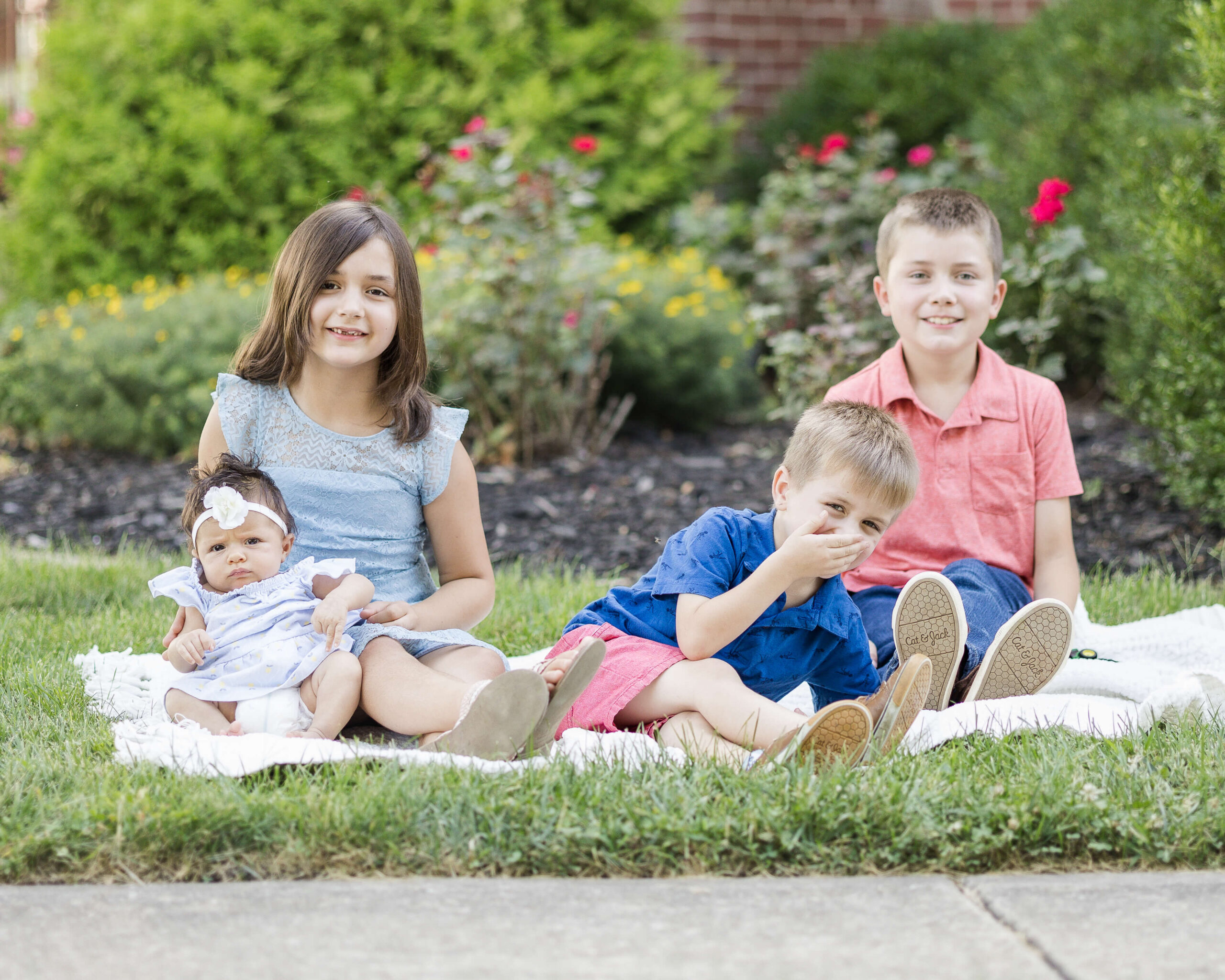 Three young siblings play on a picnic blanket in a garden with their infant sister after atttending daycares in springfield il