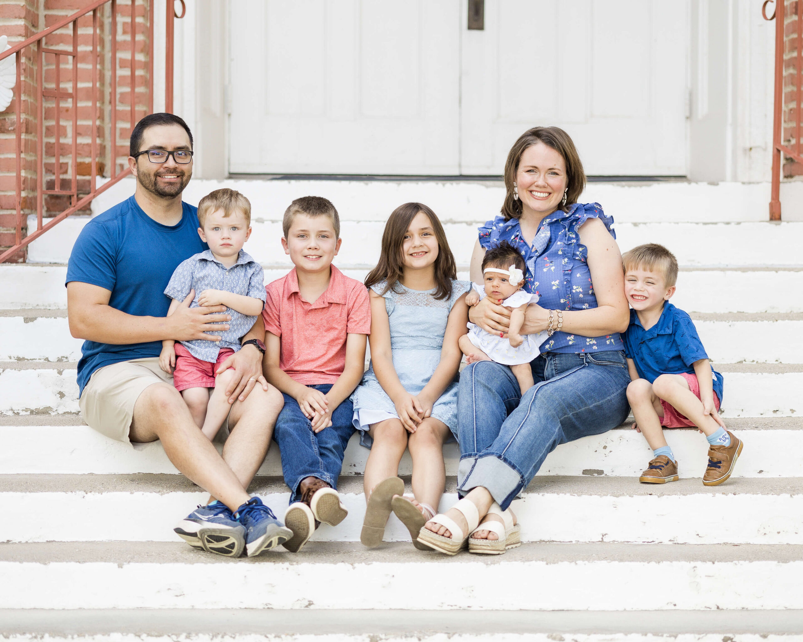 Smiling mom and dad sit on church steps in blue and pink with their four toddlers and infant girl after finding daycares in springfield il