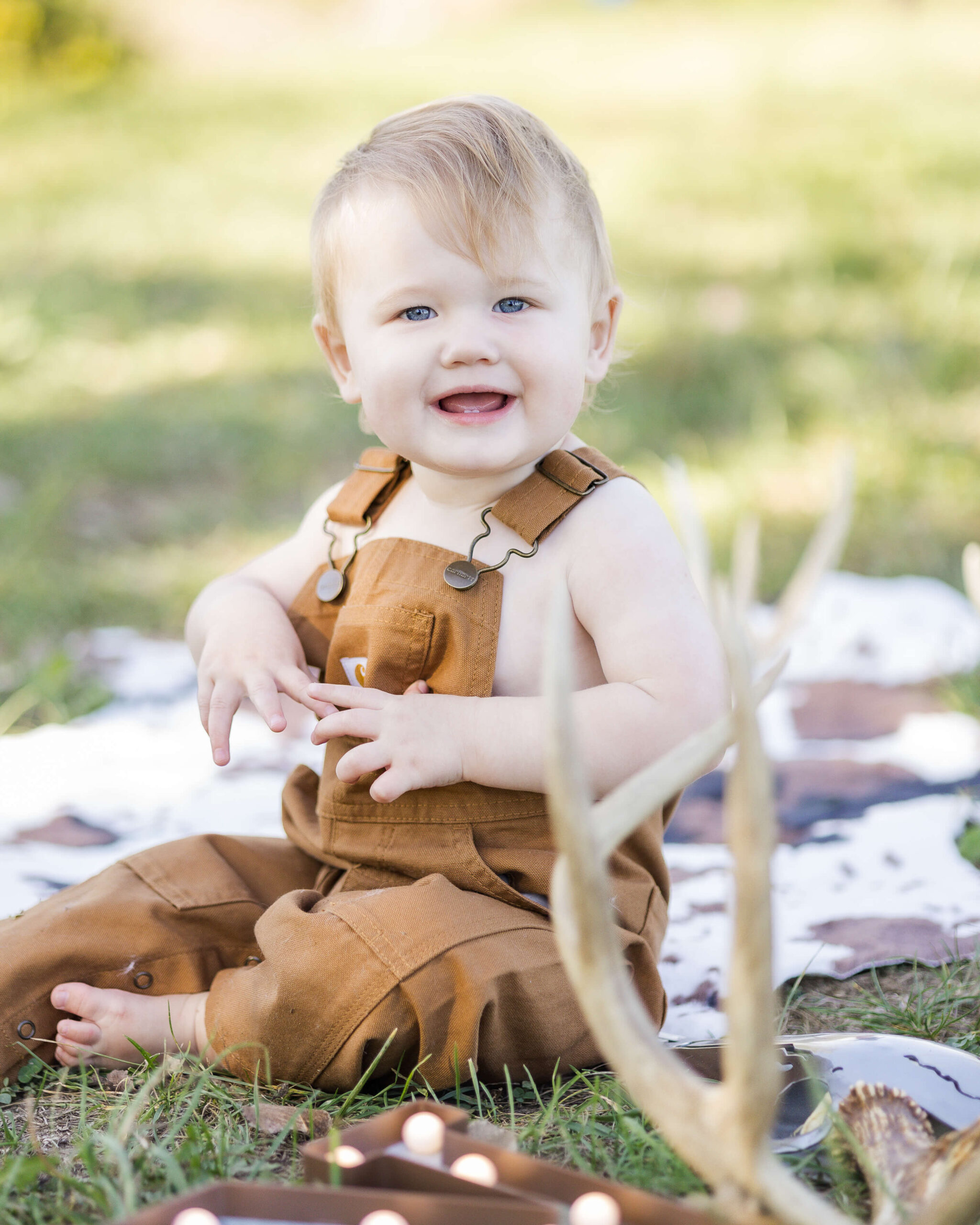 A smiling toddler in brown overalls sits in a park lawn with deer antlers