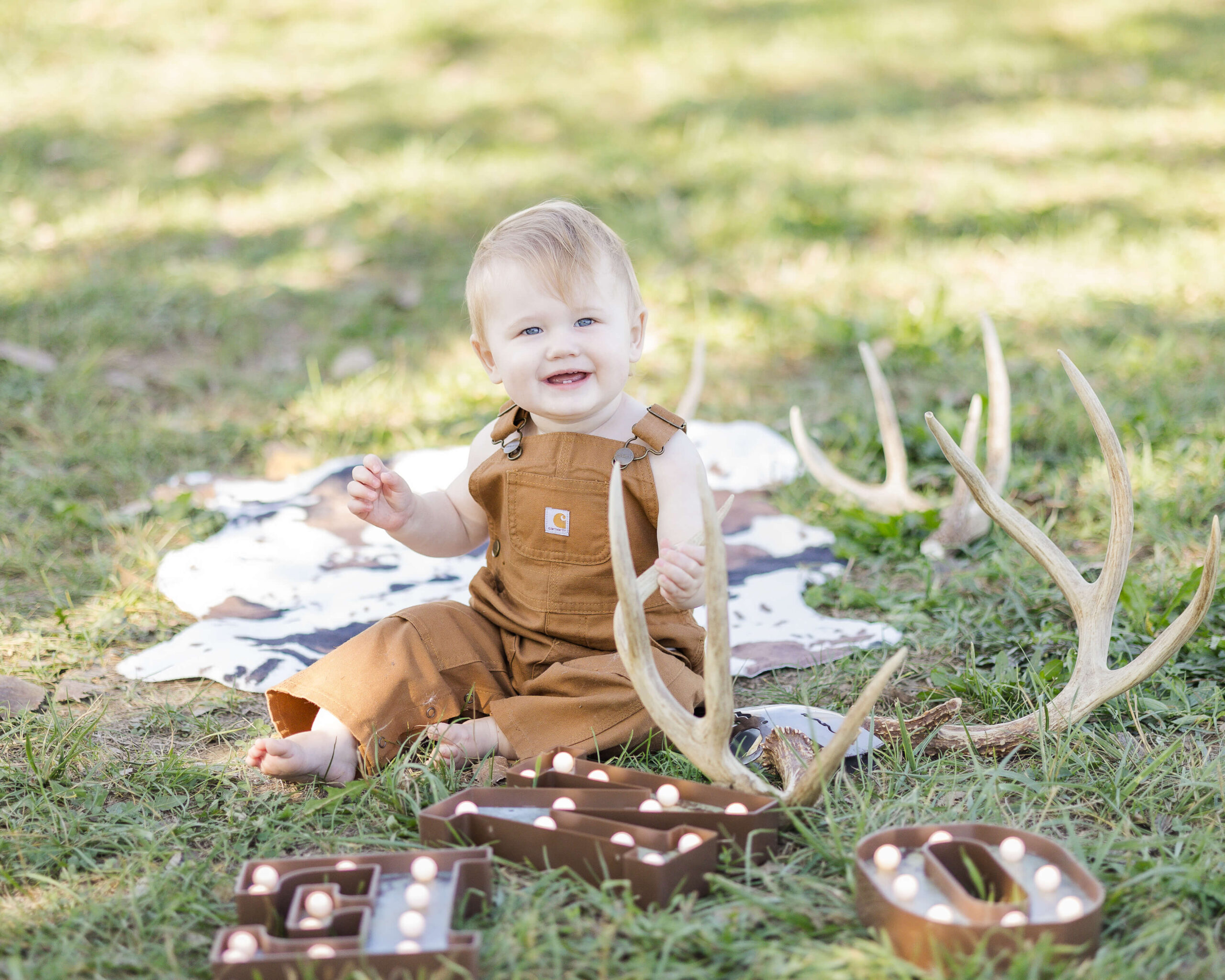 A happy toddler in brown overalls sits among deer antlers and marquee letters after visiting little lincoln's toy shop
