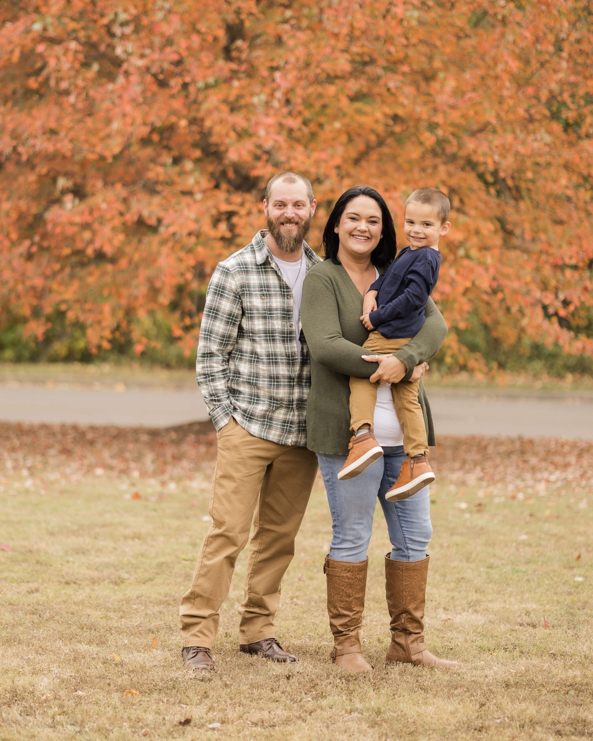 A smiling mom and dad in green and tan witht heir toddler son in mom's arms in a park before exploring music venues in springfield il