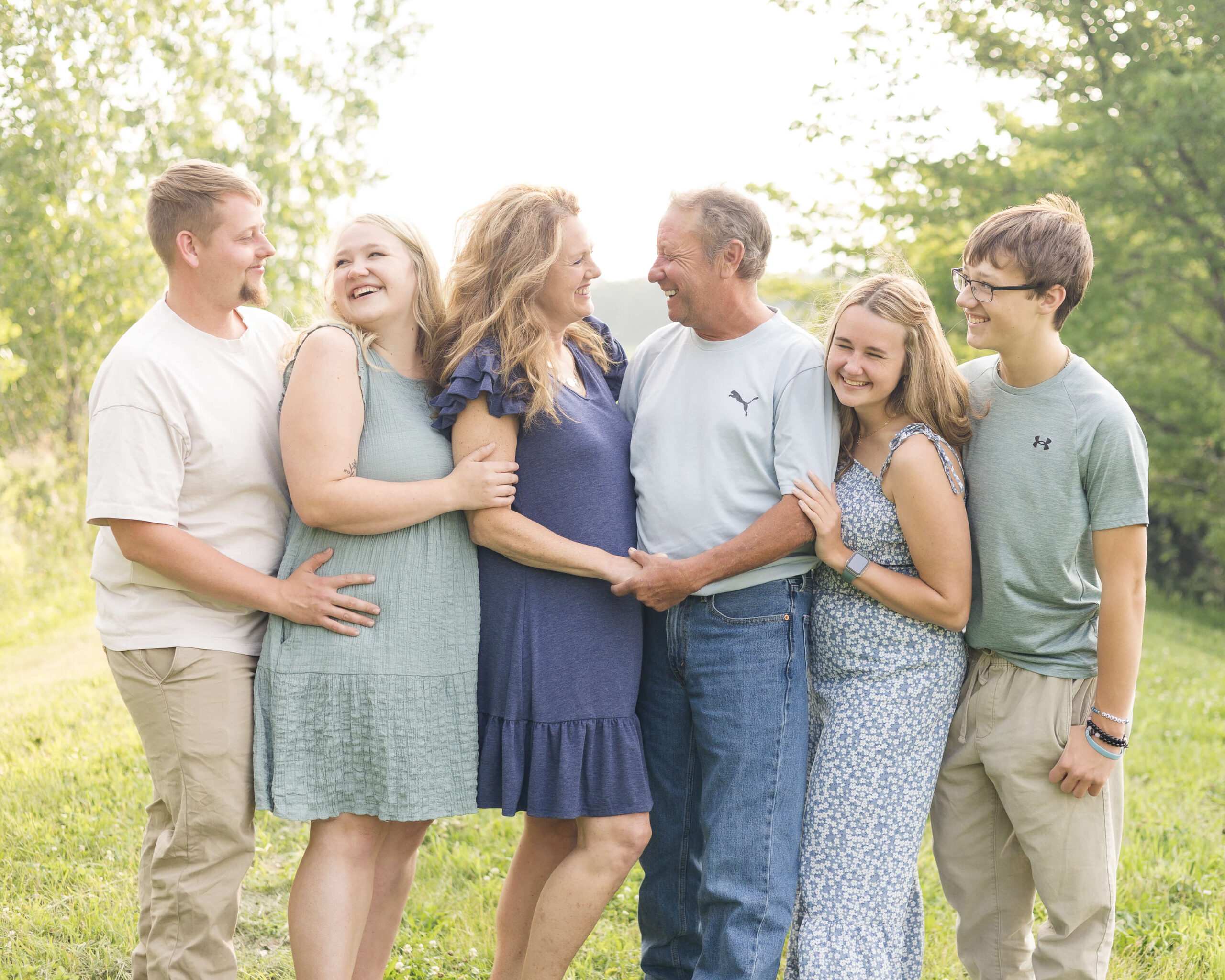A mom and dad smile at each other while surrounded by their laughing teen children in a park lawn