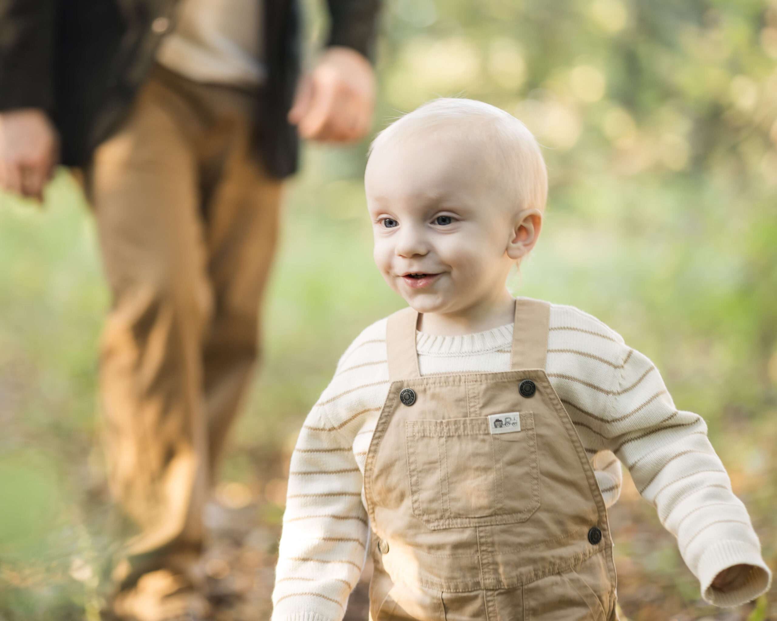 A toddler boy in tan overalls and white sweater runs in a park in front of dad after visiting children's museums in Springfield, IL