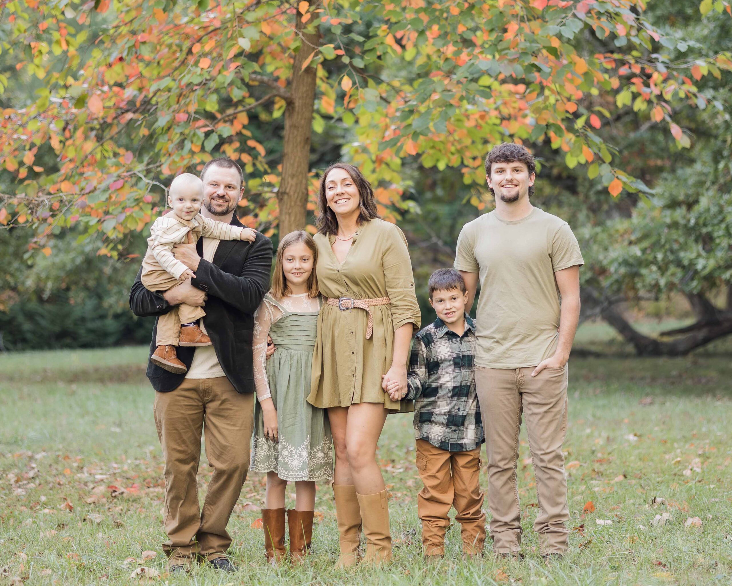 A happy mom and dad stand with their teen son and three toddlers in a park in fall