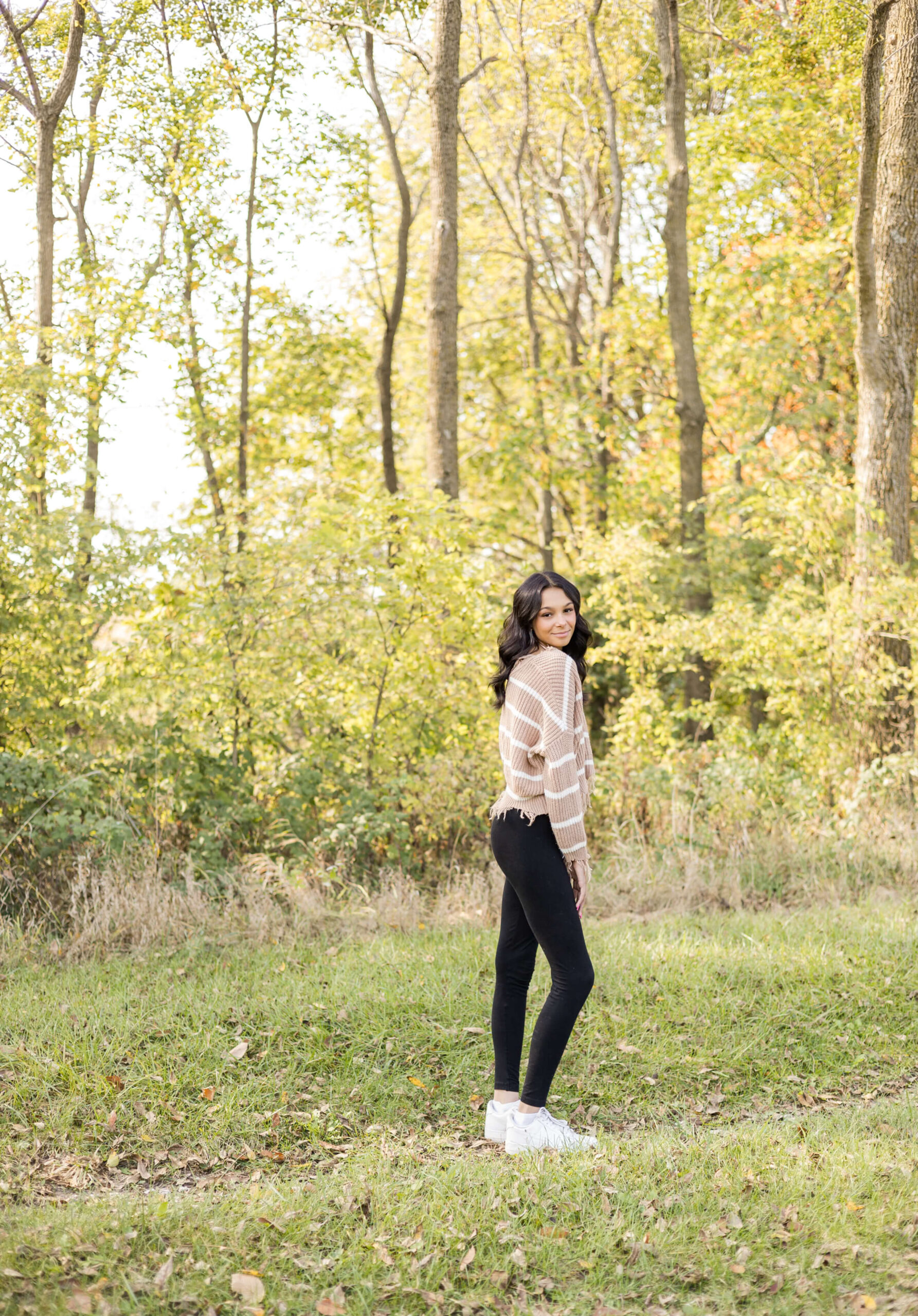 A high school senior in a tan sweater and black pants walks in a park while smiling over her shoulder after enjoying summer camps in Springfield, IL