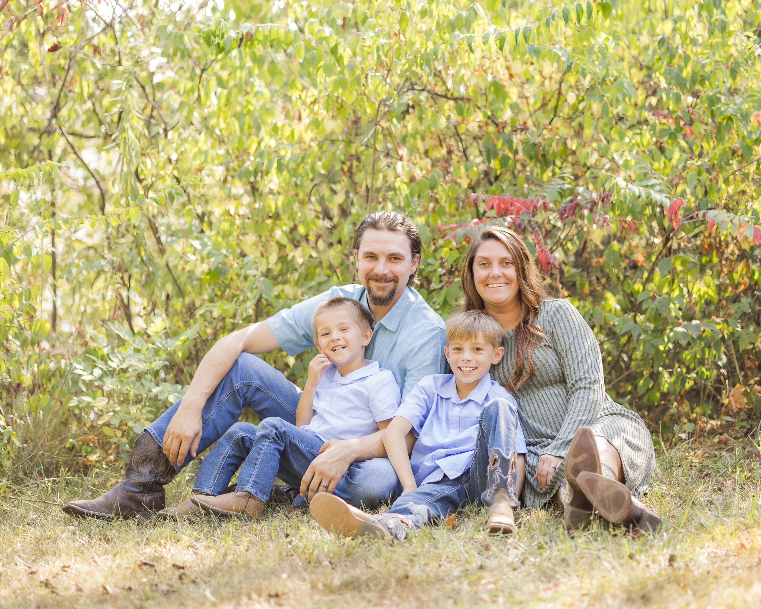 A mom and dad smile while sitting in a lawn with their toddler sons in their laps