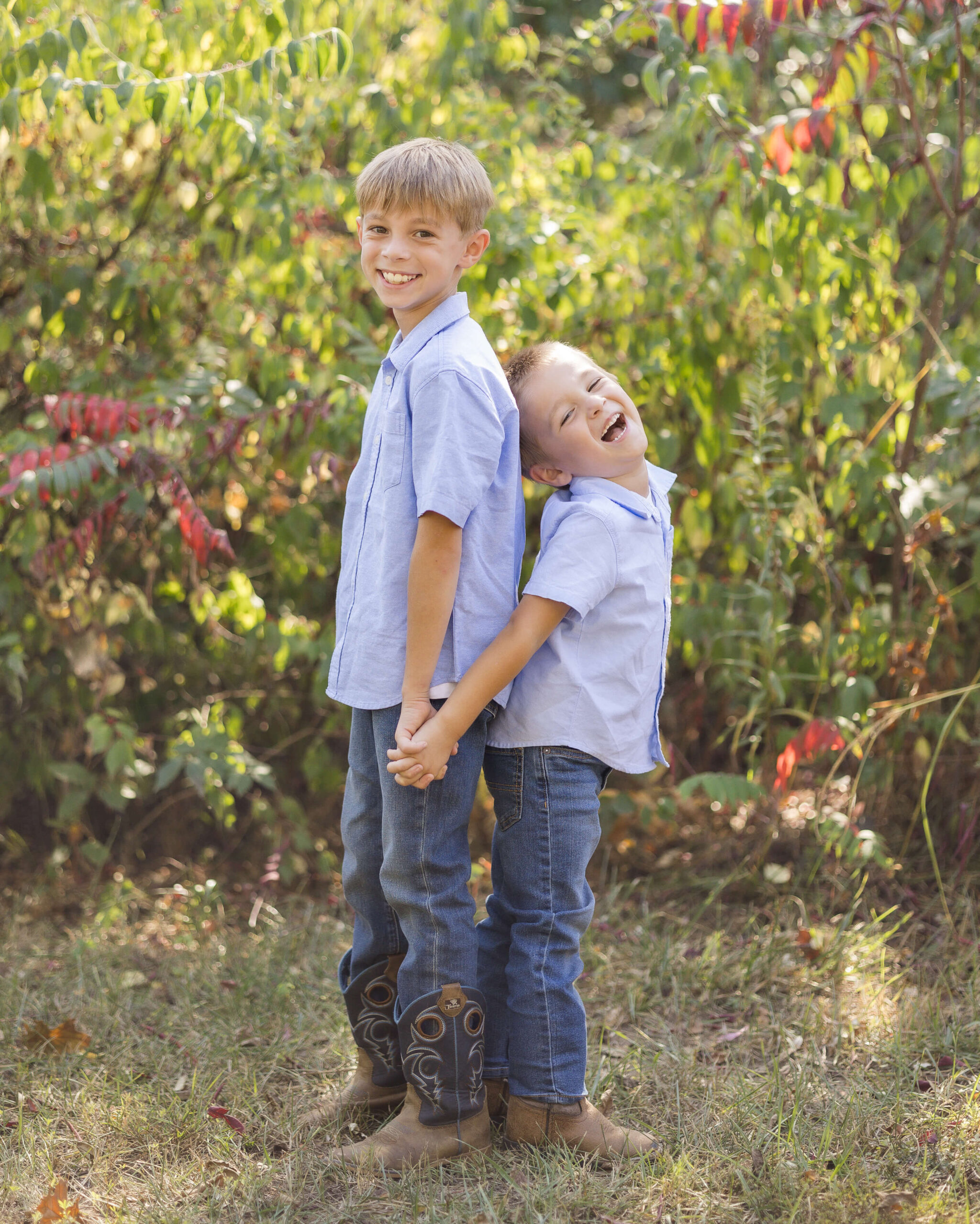 Happy toddler brothers in matching blue shirts and jeans stand back to back holding hands in a park before some summer camps in Springfield, IL