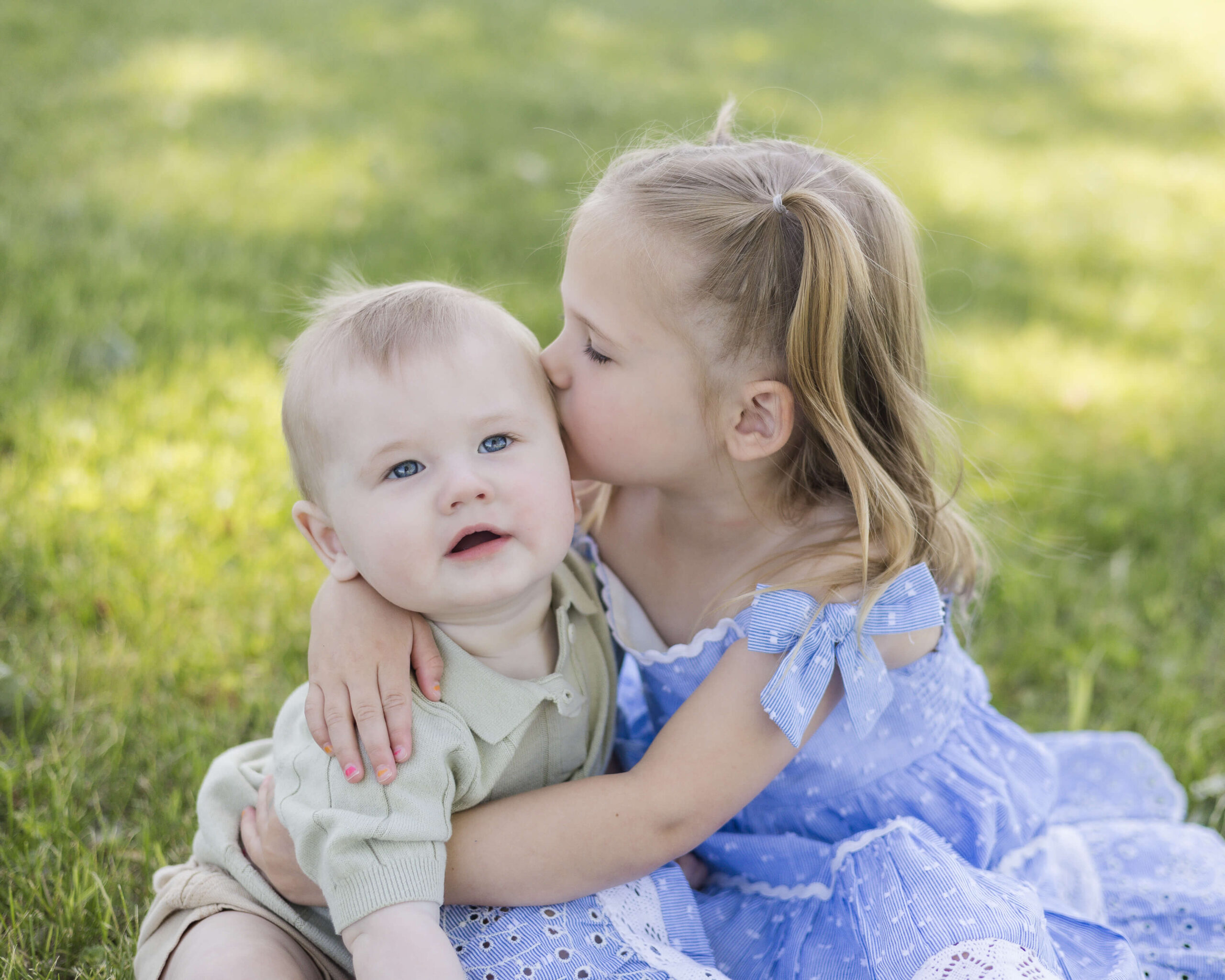 A toddler girl in a blue dress kisses the cheek of her baby brother as she hugs him sitting in a lawn