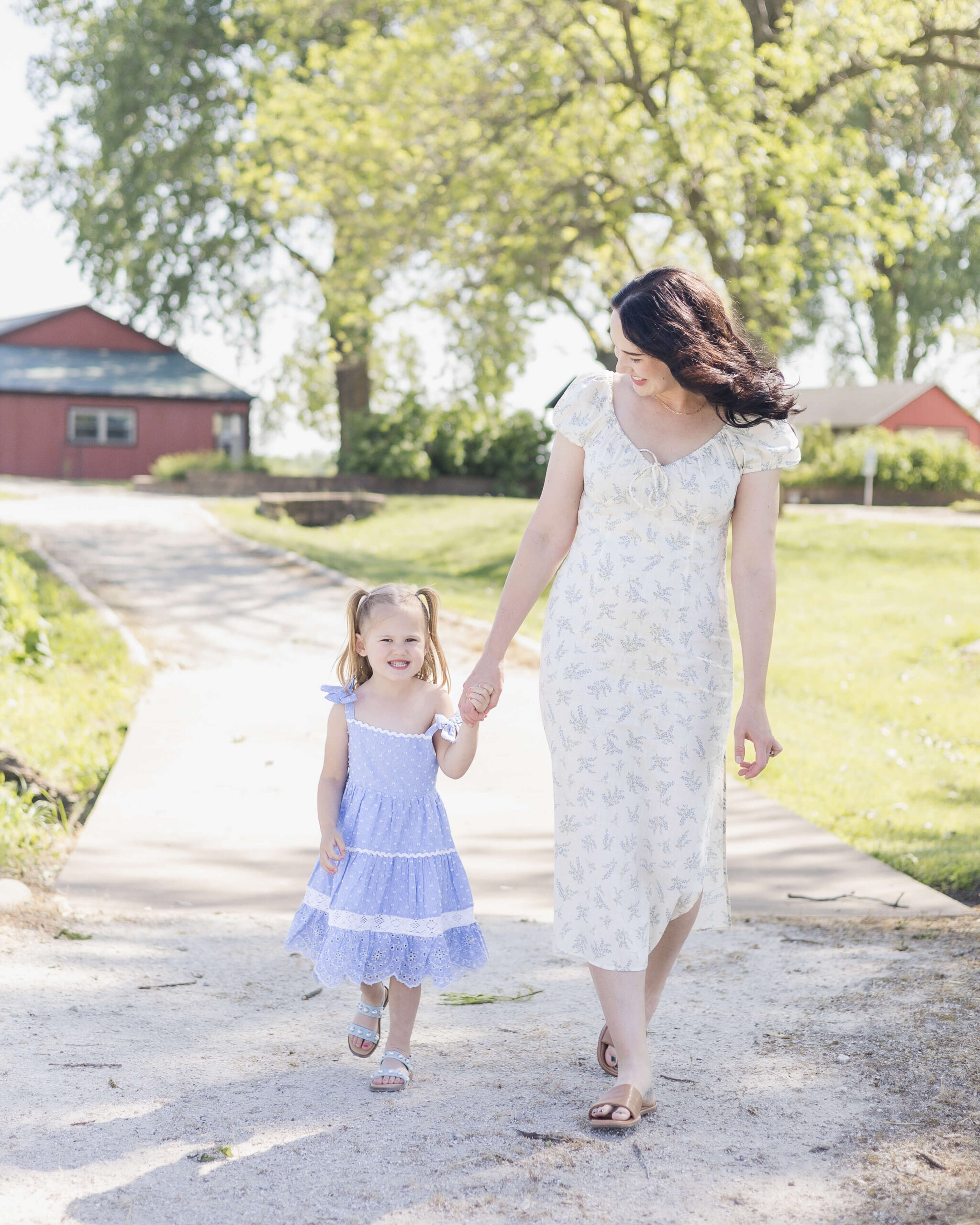 A smiling mom in a white dress walks a park sidewalk holding hands with her toddler daughter in a blue dress after some swim lessons in Springfield, IL