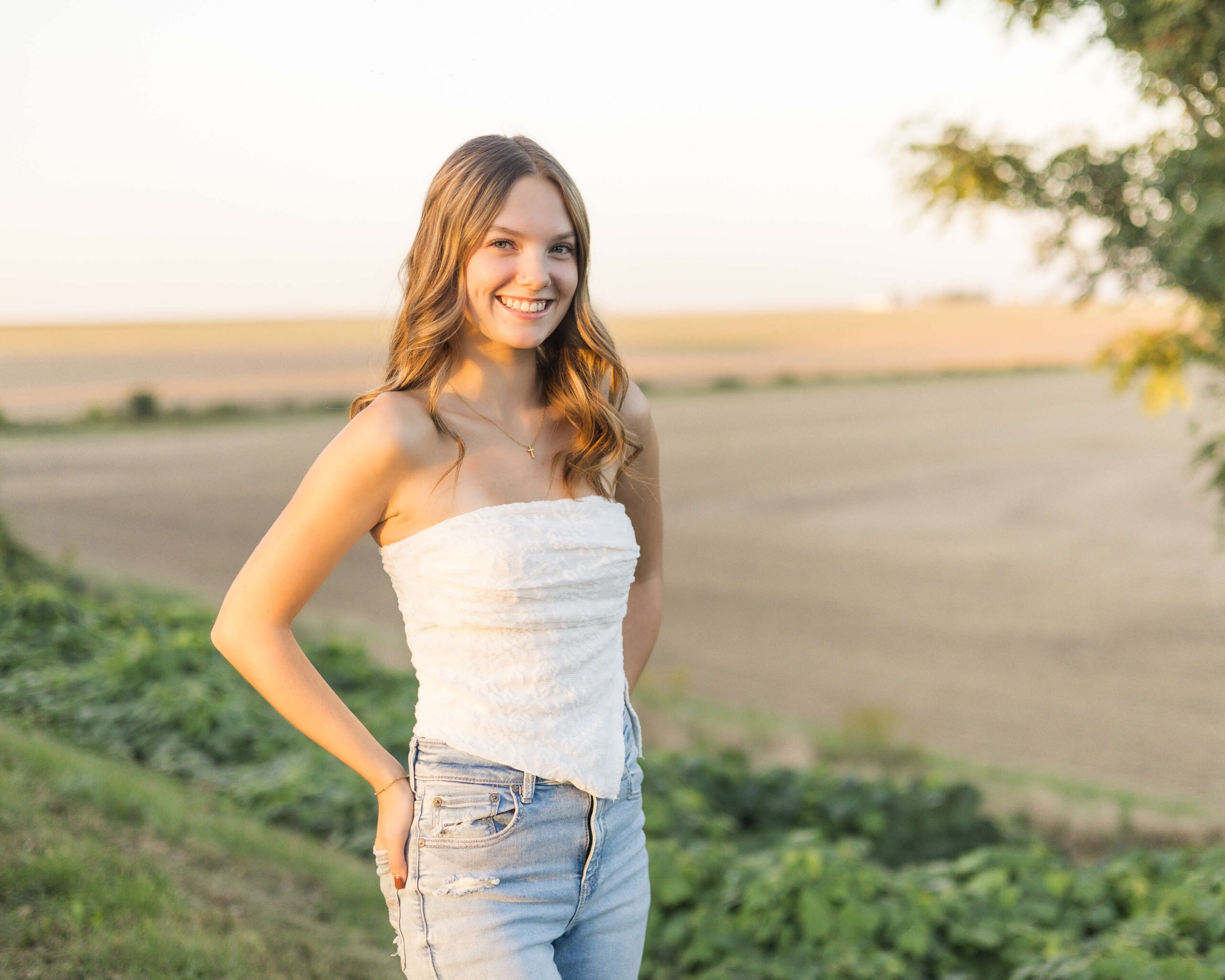 A happy high school senior smiles in a white shirt and jeans at sunset overlooking a farm