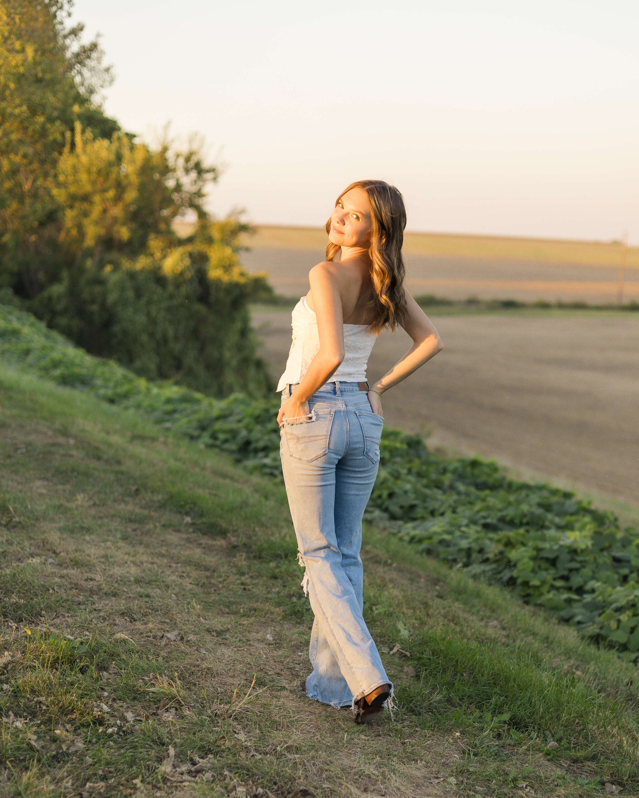 A smiling high school senior walks on a trail overlooking a farm at sunset with hands in her back jean pockets after visiting tanning salons in Springfield, IL